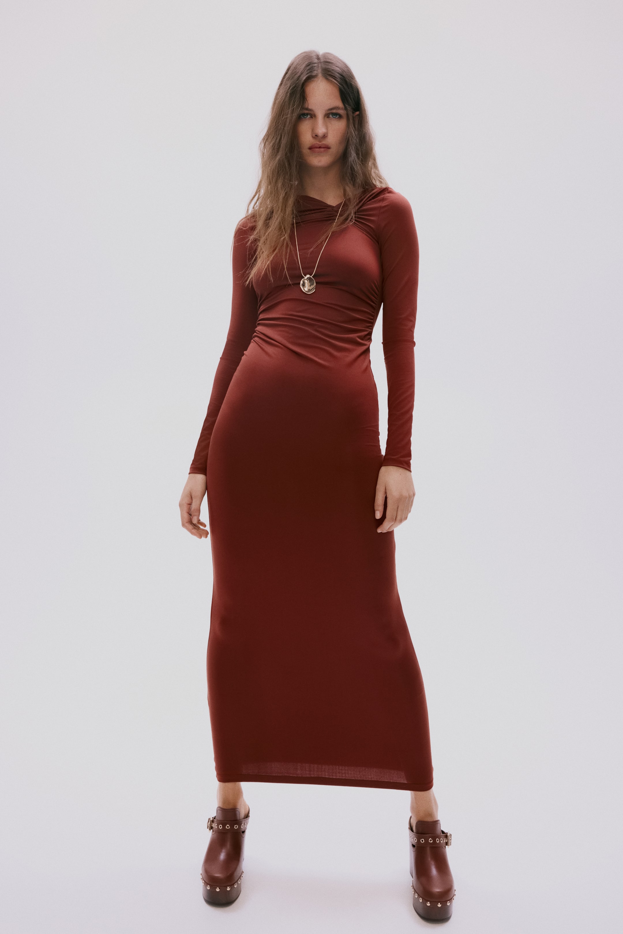 View larger image: A rust red maxi dress is presented front-on, featuring a draped cowl neckline and long sleeves. It showcases a body-skimming fit with ruching details along the torso.