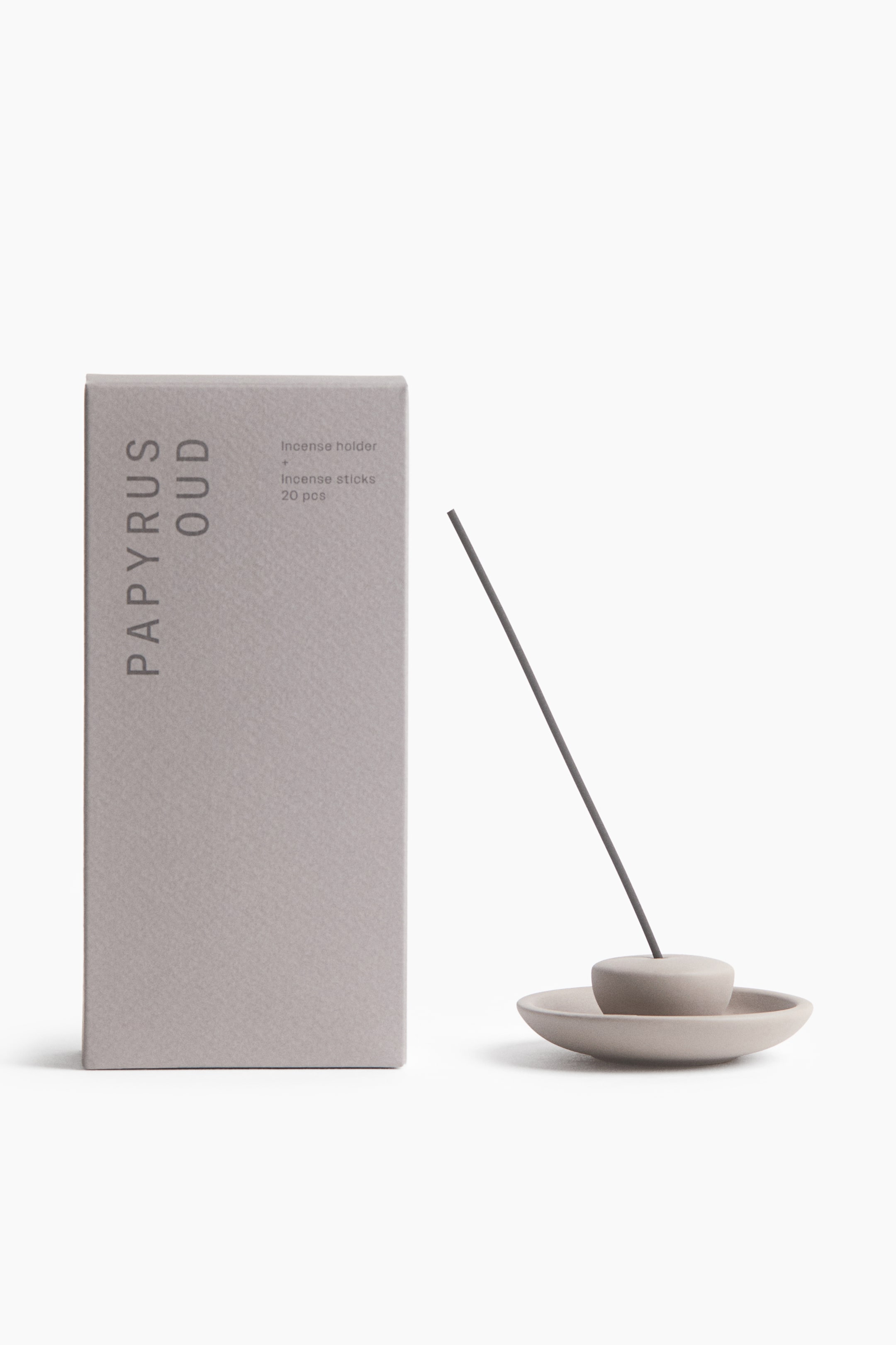View larger image: An incense holder in matt grey stoneware, featuring a shallow round dish and a small rounded central holder, is holding a single grey incense stick at an angle. A matching rectangular grey packaging box printed with "PAPYRUS OUD" is standing to the left of the holder.