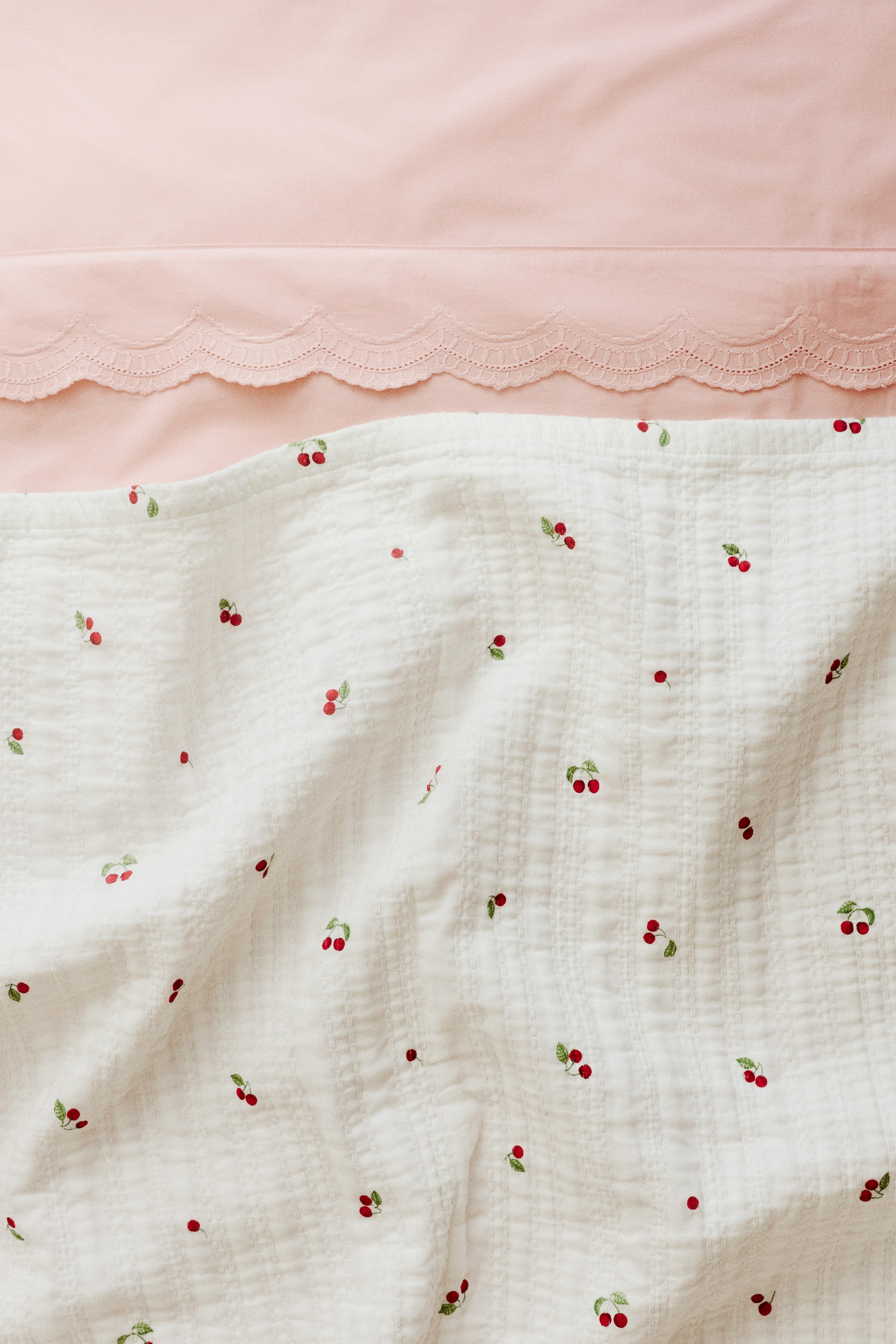 View larger image: A white soft cotton muslin bedspread is showcasing its front. It features an all-over print of red cherries with green stems, and visible stitching creates a textured surface.