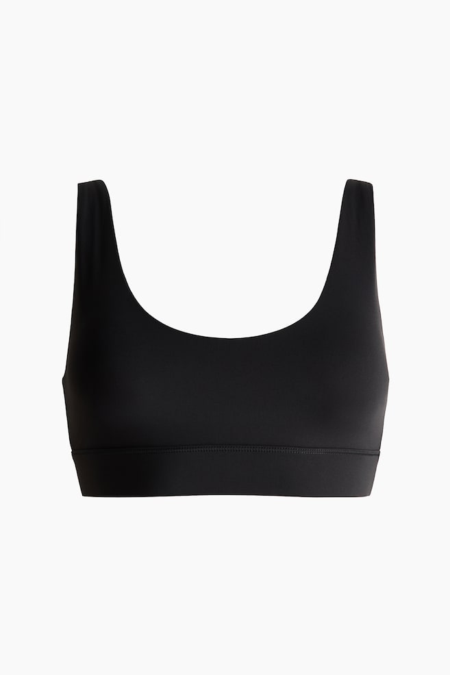 A black sports bra is shown from the front, highlighting a scoop neckline and broad tank straps. A wide band with prominent stitching forms the bottom hem. - 1