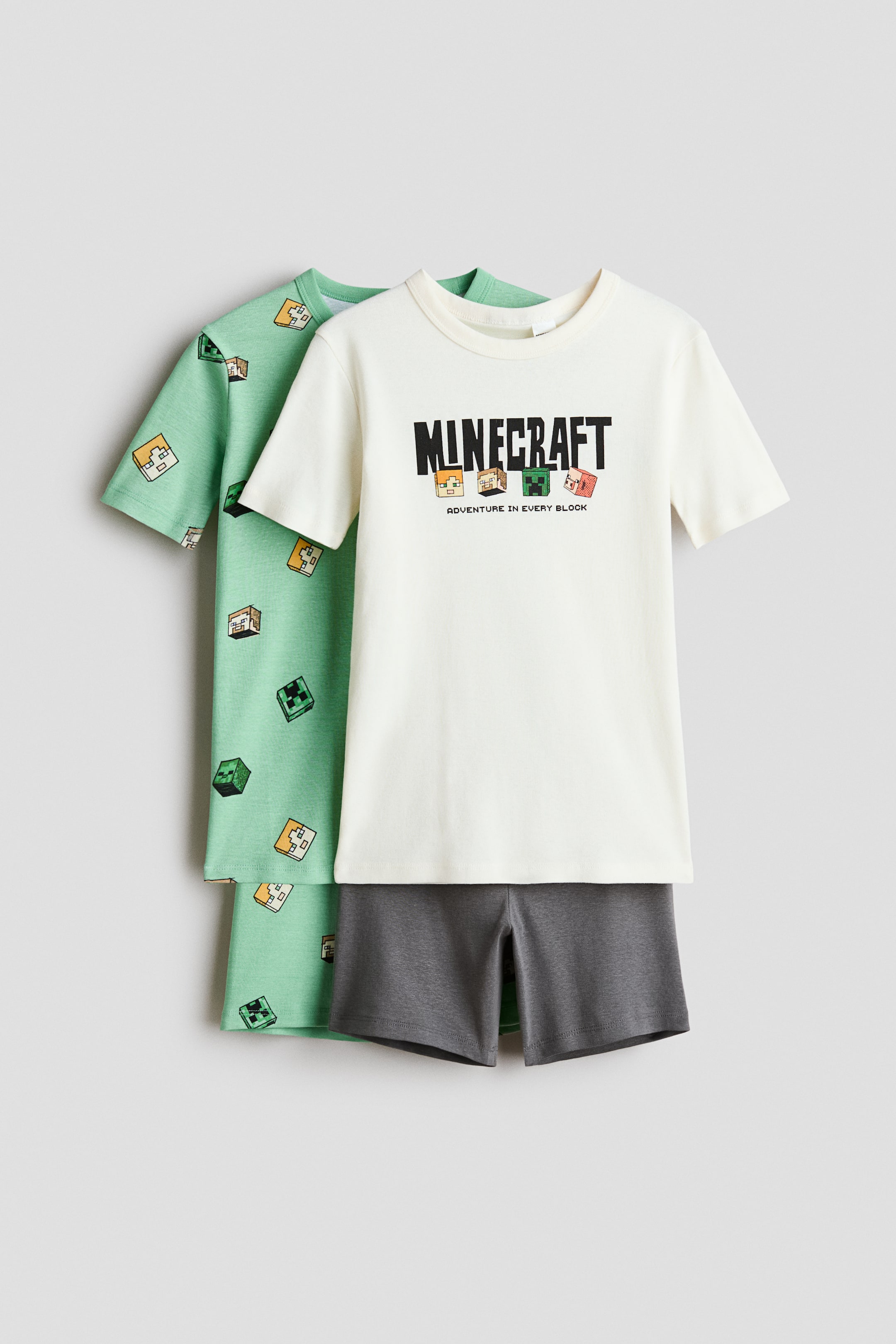 View larger image: The children's Minecraft pajama set includes an off-white short-sleeve t-shirt featuring a front-facing "MINECRAFT ADVENTURE IN EVERY BLOCK" graphic. It's layered over a light green short-sleeve t-shirt displaying an all-over pattern of various Minecraft blocks. The set is completed with dark gray shorts.