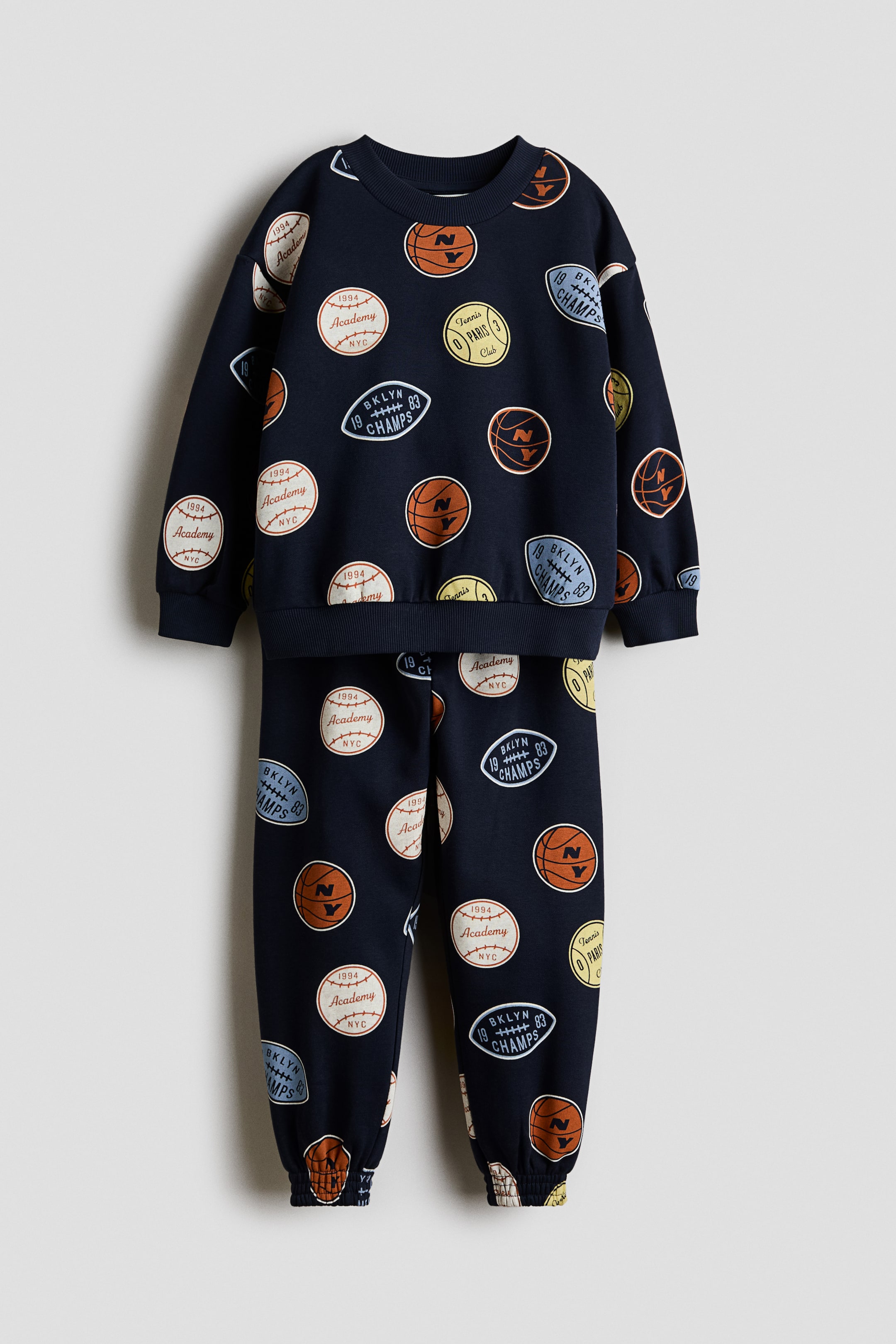 View larger image: A navy blue two-piece tracksuit is displayed front-facing, featuring a crew-neck sweatshirt paired with matching joggers. The sweatshirt boasts long sleeves, ribbed cuffs, and a ribbed hem, while the joggers are crafted with ribbed ankle cuffs. Both items are adorned with an all-over sports-themed patch pattern, showcasing basketballs, baseballs, footballs, and tennis club emblems in tan, brown, white, blue, and yellow hues.
