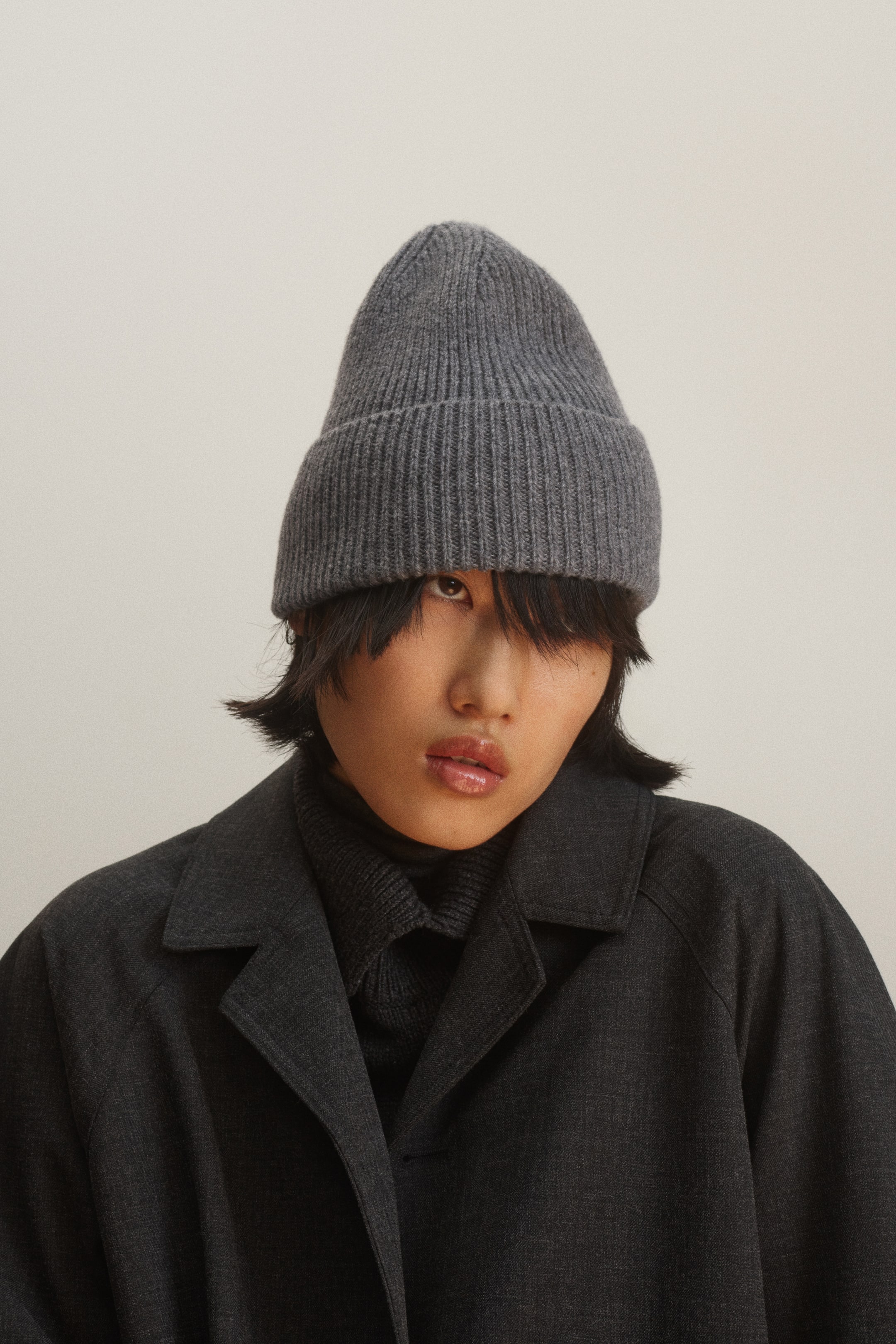 View larger image: A grey beanie in a soft rib-knit featuring a wide turn-up hem is being worn by the model. The beanie is styled with a dark grey collared coat layered over a matching high-neck top.