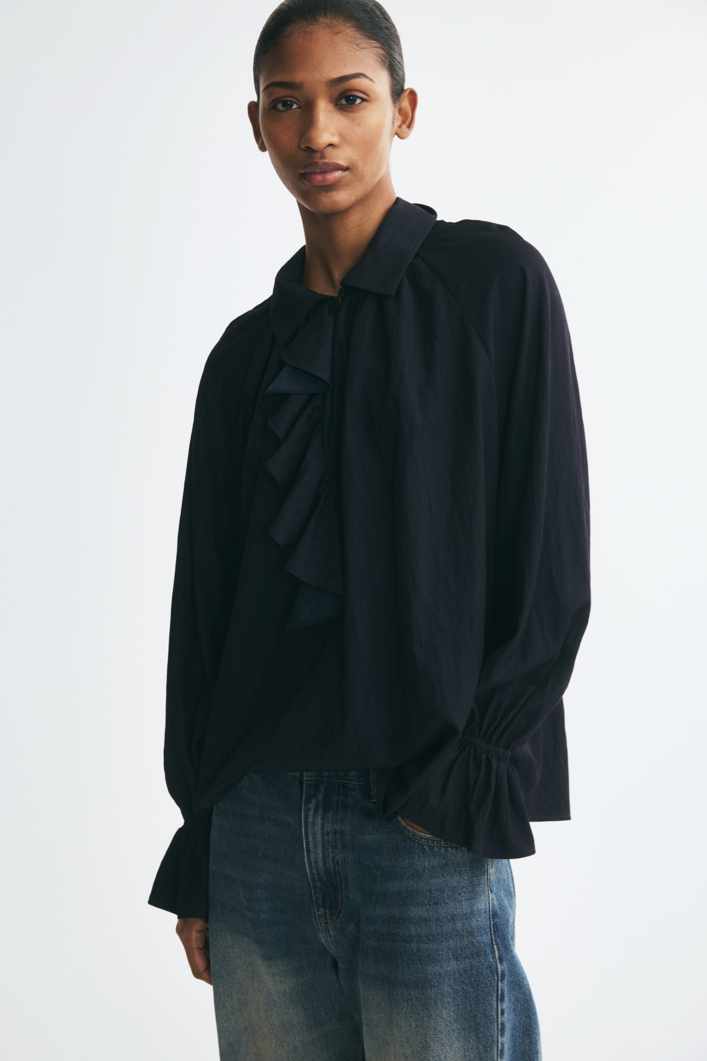 This dark navy blue blouse is shown from the front, featuring a pointed collar and a cascading ruffle detail down the center placket. The garment has a relaxed, gathered silhouette with long sleeves ending in ruffled cuffs.