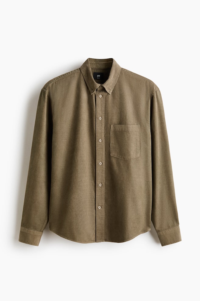 Una camisa de pana verde oliva orientada al frente presenta un cuello con botones y una tapeta completa de botones en la parte delantera. Incluye un único bolsillo abierto en el pecho a la izquierda, mangas largas con puños abotonados y un dobladillo redondeado. - 1