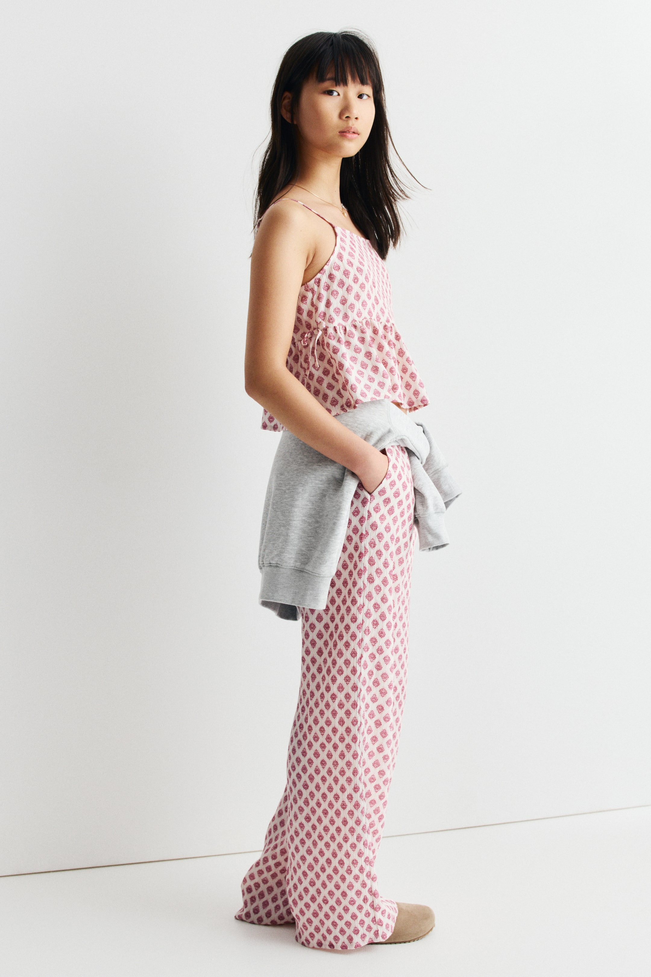 Ver imagen más grande: Un conjunto de dos piezas en color rosa claro, que consta de un top corto tipo camisola con tirantes finos y un dobladillo fruncido, y pantalones anchos a juego, se ve desde el lado. Ambas piezas presentan un patrón de motivos repetidos en rosa oscuro, y una sudadera gris está atada alrededor de la cintura.