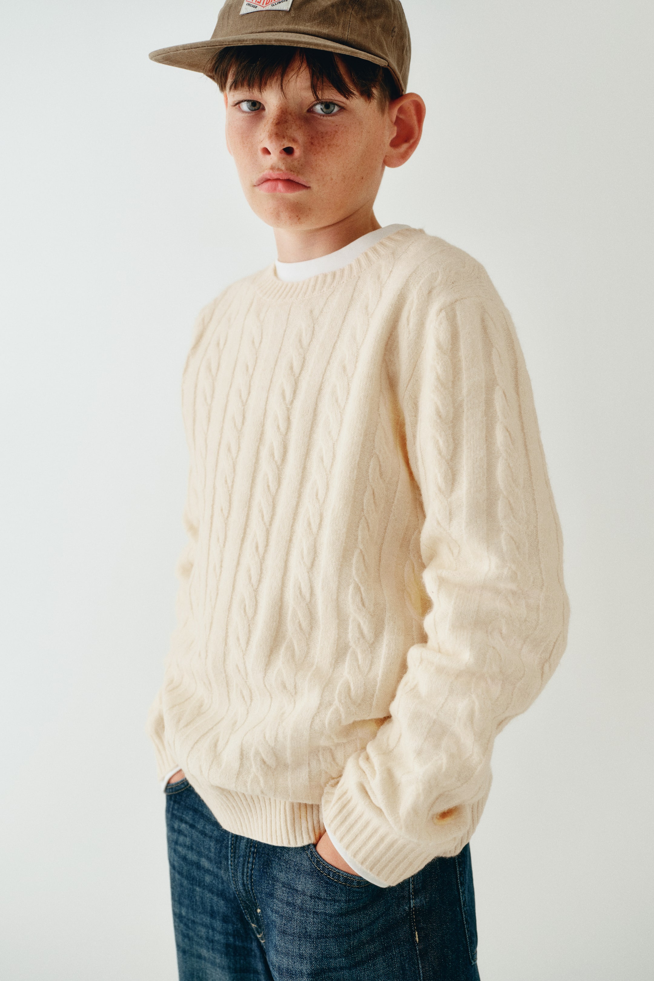 Agrandir l'image: Un pull en maille torsadée de couleur crème est présenté de face, mettant en valeur un col rond côtelé, des épaules tombantes et des finitions côtelées aux poignets et à l’ourlet. Le pull est porté en superposition sur un t-shirt blanc et assorti à un jean bleu.