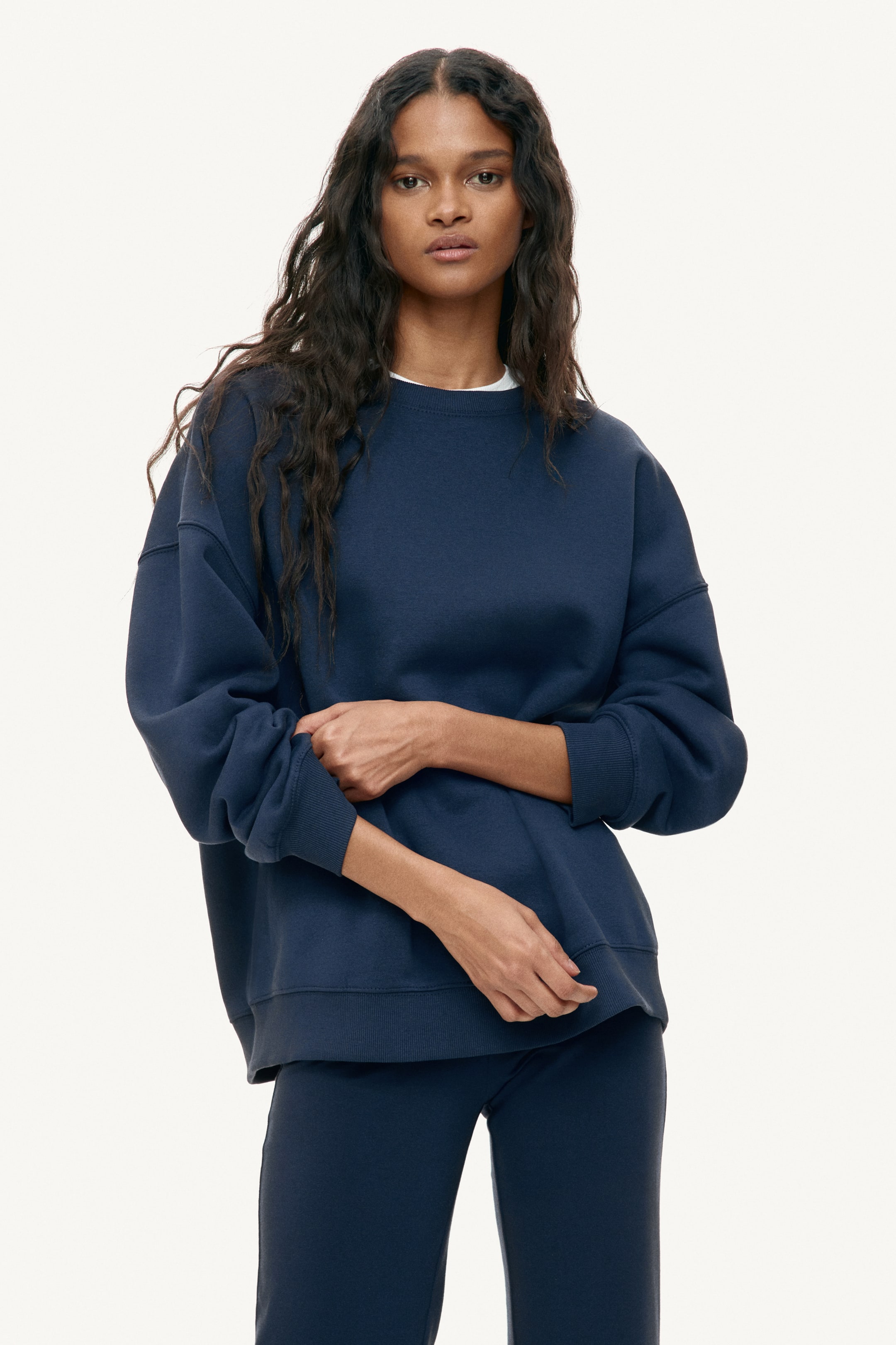 View larger image: Discover our navy blue oversized crew neck sweatshirt, showcasing long sleeves with ribbed cuffs and a ribbed hem. A white t-shirt subtly peeks out at the neckline, adding a touch of contrast. Complementing the look, matching navy blue pants complete the set, perfect for a cohesive and stylish ensemble.