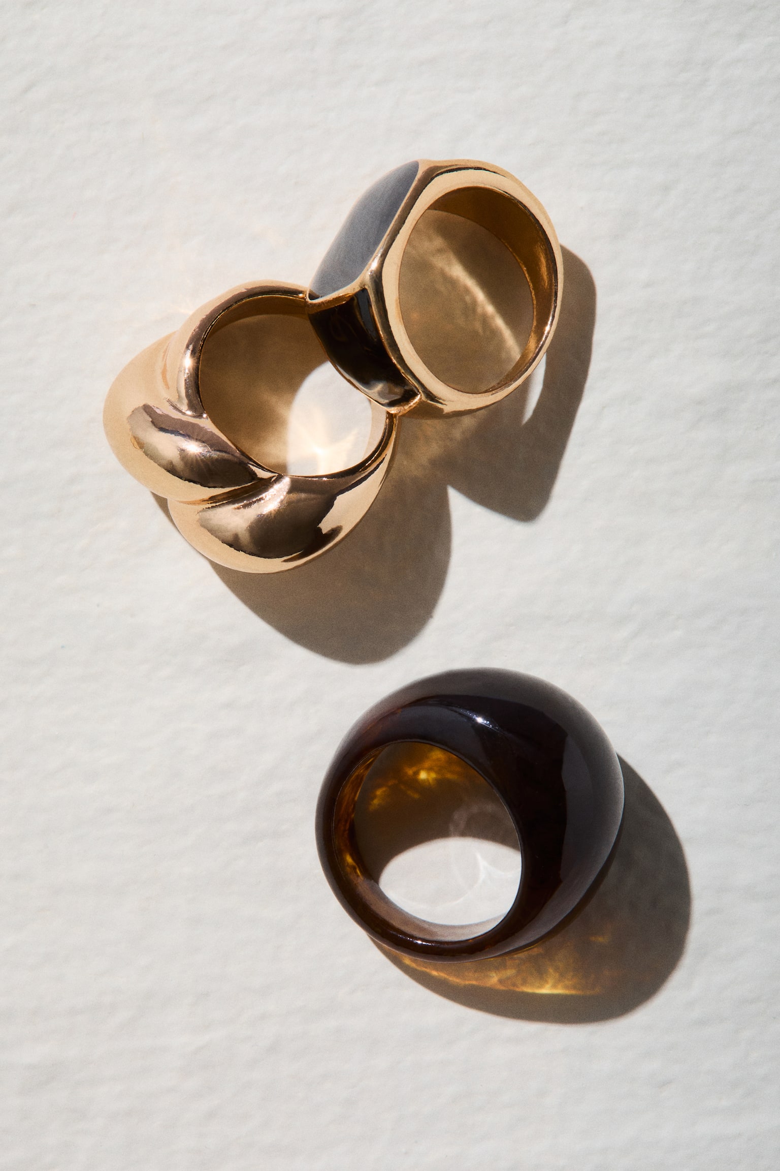 Three chunky rings boast various designs, each angled slightly to the right. On the left sits a rounded tortoiseshell resin ring. Centrally placed is a twisted gold-toned metal ring, while to the right, a gold-toned metal signet ring features a rectangular black enamel top. - 1