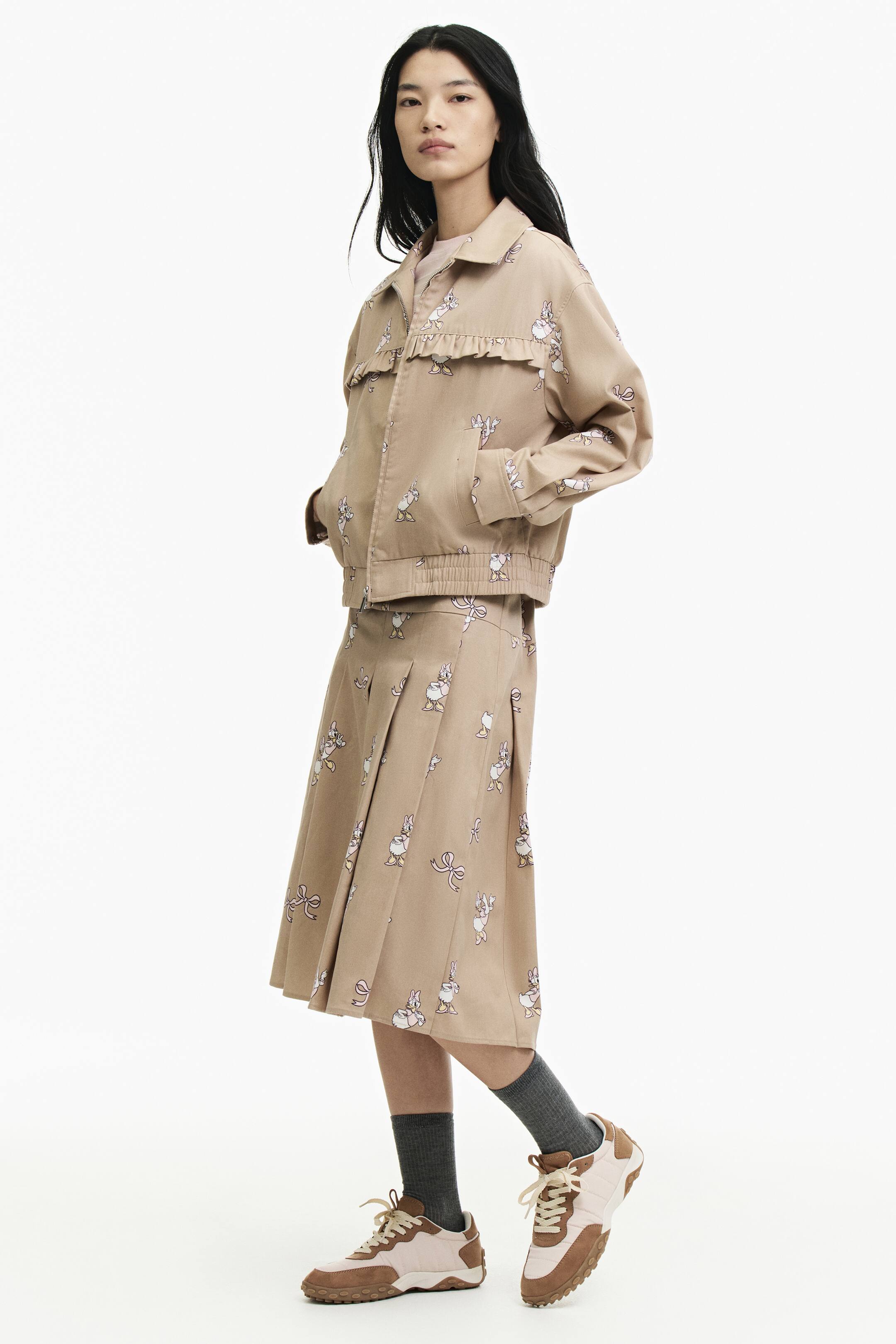 Xem ảnh lớn hơn: Một chiếc áo khoác cotton twill màu be với họa tiết hoạt hình phủ toàn bộ đang được người mẫu mặc, tay đặt trong túi chéo phía trước. Áo khoác có cổ áo, cầu vai ngực viền bèo nhún và gấu áo chun, được phối cùng chân váy xếp ly in họa tiết đồng bộ.