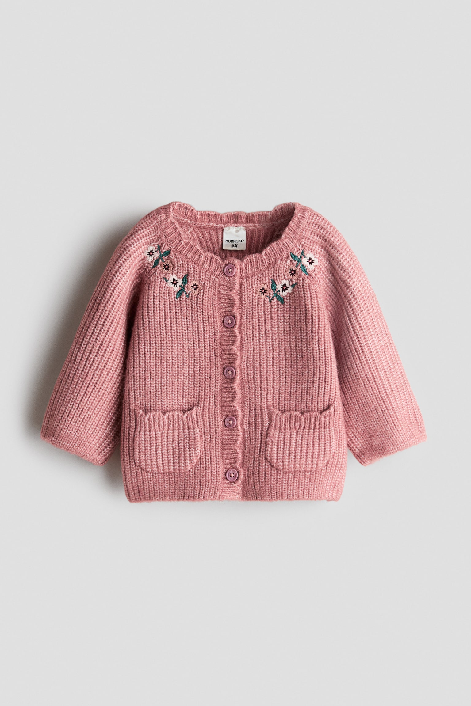 A pink, soft English rib-knit cardigan is shown from the front, featuring delicate floral embroidery near the shoulders and a round, scalloped neckline. It is detailed with a button-down front and two small, open front pockets with matching scalloped edges.
