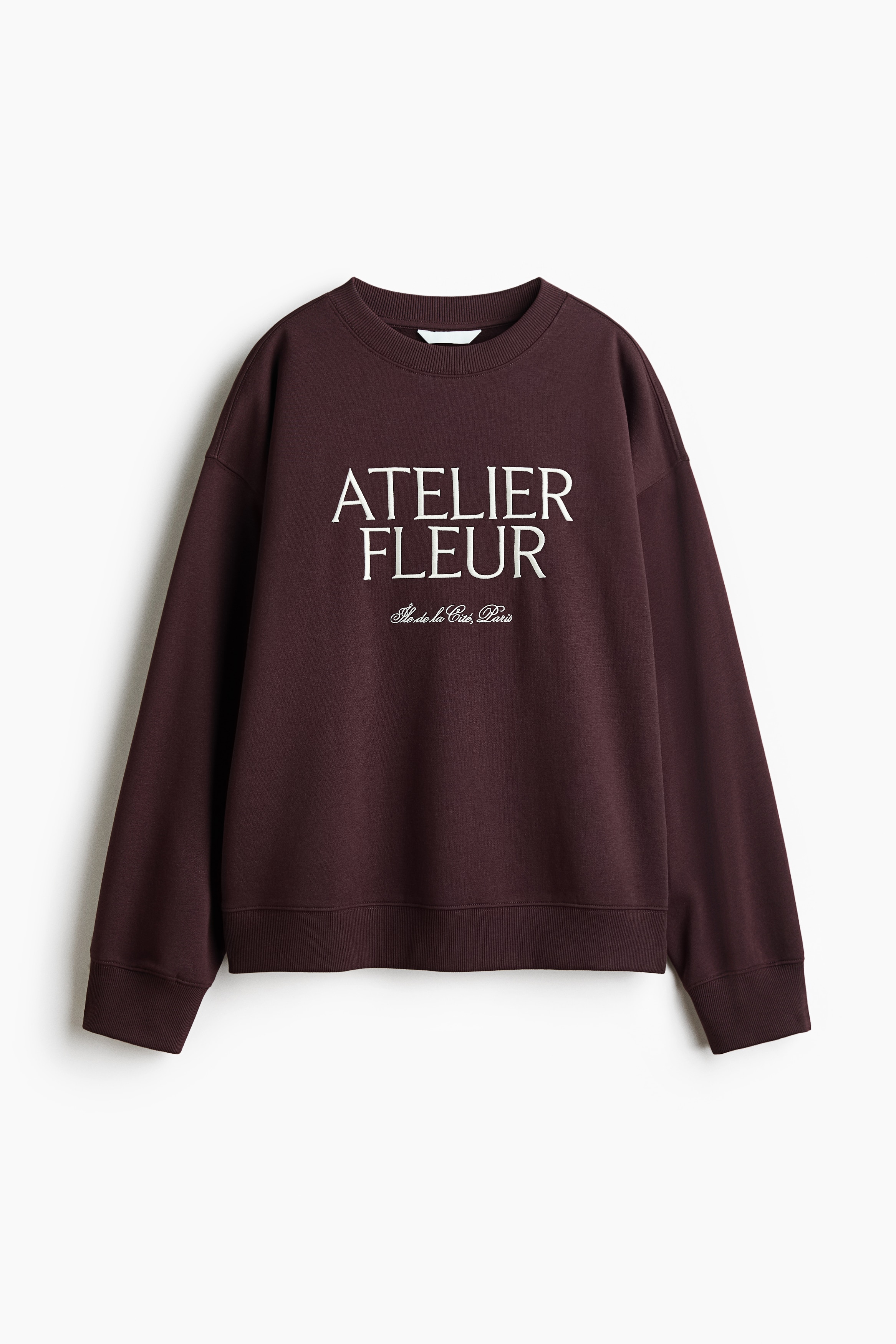 A dark burgundy, oversized sweatshirt featuring white text reading "ATELIER FLEUR" on the front is being worn by the model. The sweatshirt features a ribbed crew neckline and dropped shoulders, and is layered over a white t-shirt and styled with blue jeans.