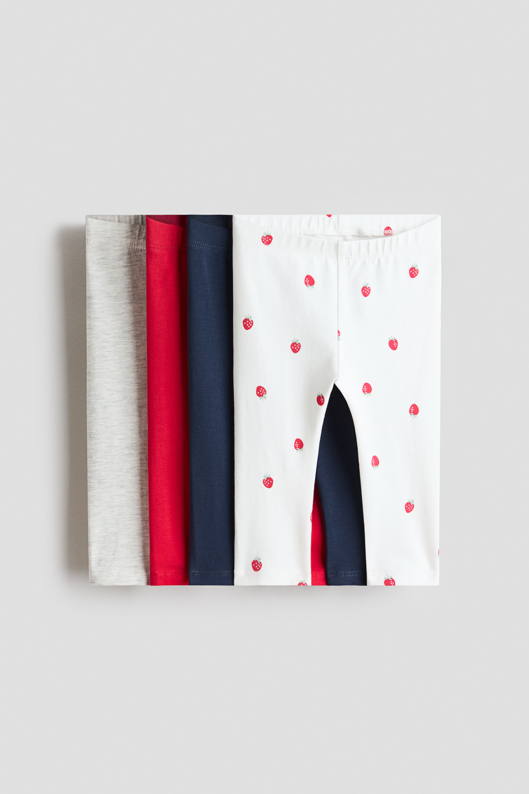 Ingrandisci l'immagine: I leggings sono presentati come una pila piegata di quattro paia, con il paio frontale rivolto verso il FRONTE. Questo paio bianco è decorato con fragole rosse e presenta una vita con elastico coperto, mentre dietro si vedono paia tinta unita in rosso, blu navy e grigio chiaro.