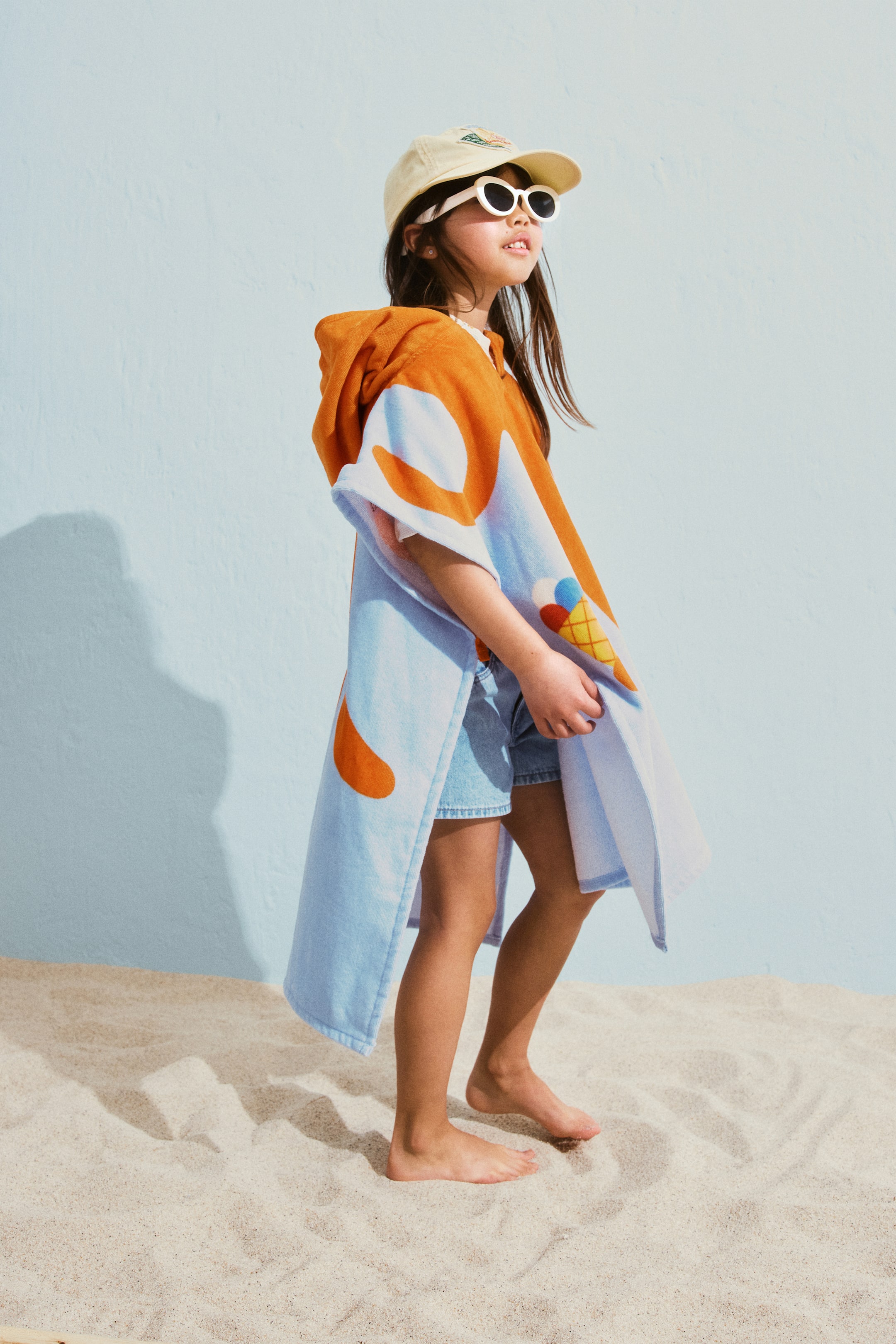이미지 확대 보기: 타월 케이프는 연한 파란색 바디에 큰 추상적인 주황색 모양과 다채로운 아이스크림 콘 그래픽이 특징이며, 주황색 후드가 디자인을 완성하고 있습니다.