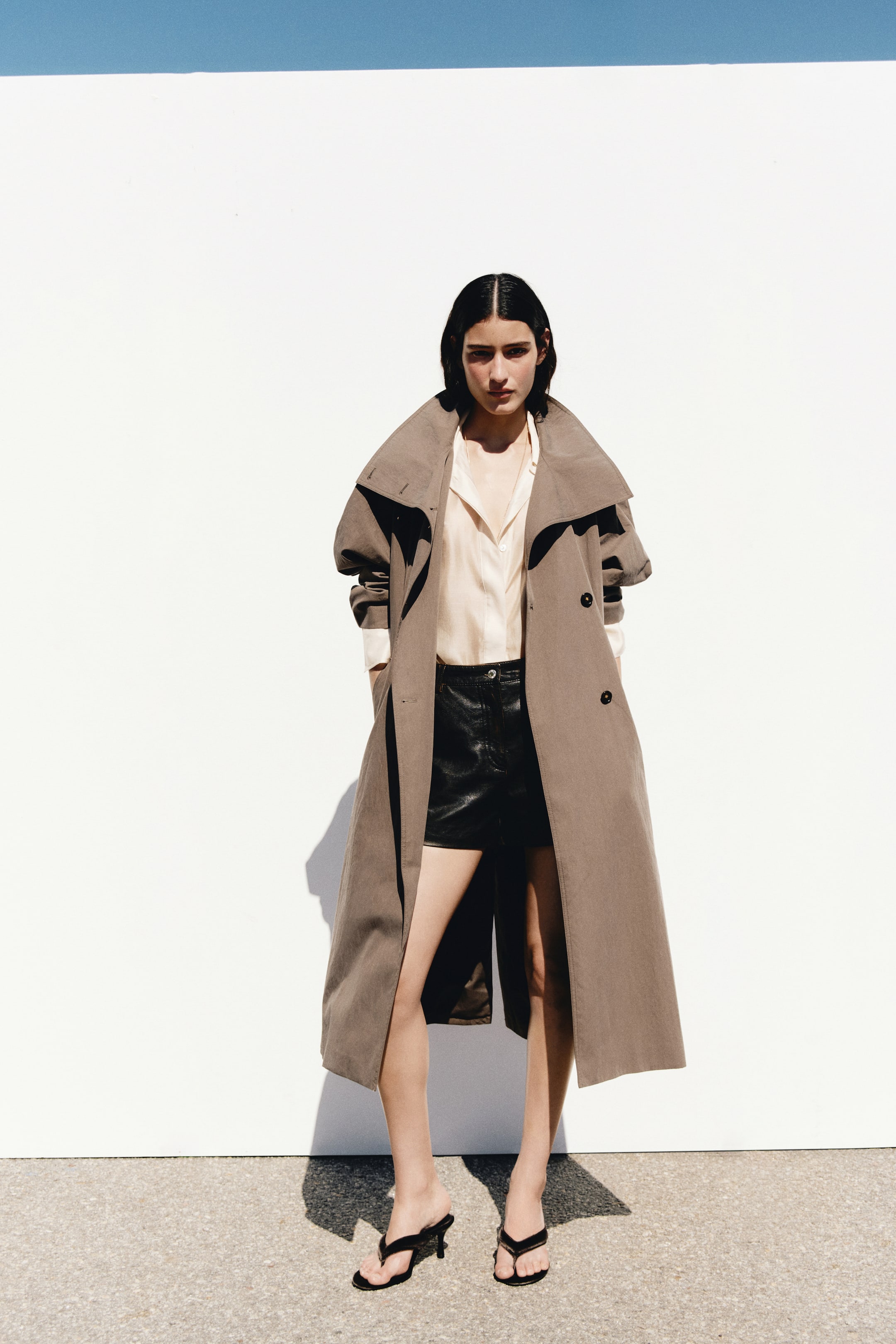 View larger image: An oversized, double-breasted taupe trench coat featuring a wide collar and front buttons is worn open by the model. The mid-length coat is layered over a light-coloured button-up shirt and black shorts, with the model standing with hands placed in the coat's pockets.