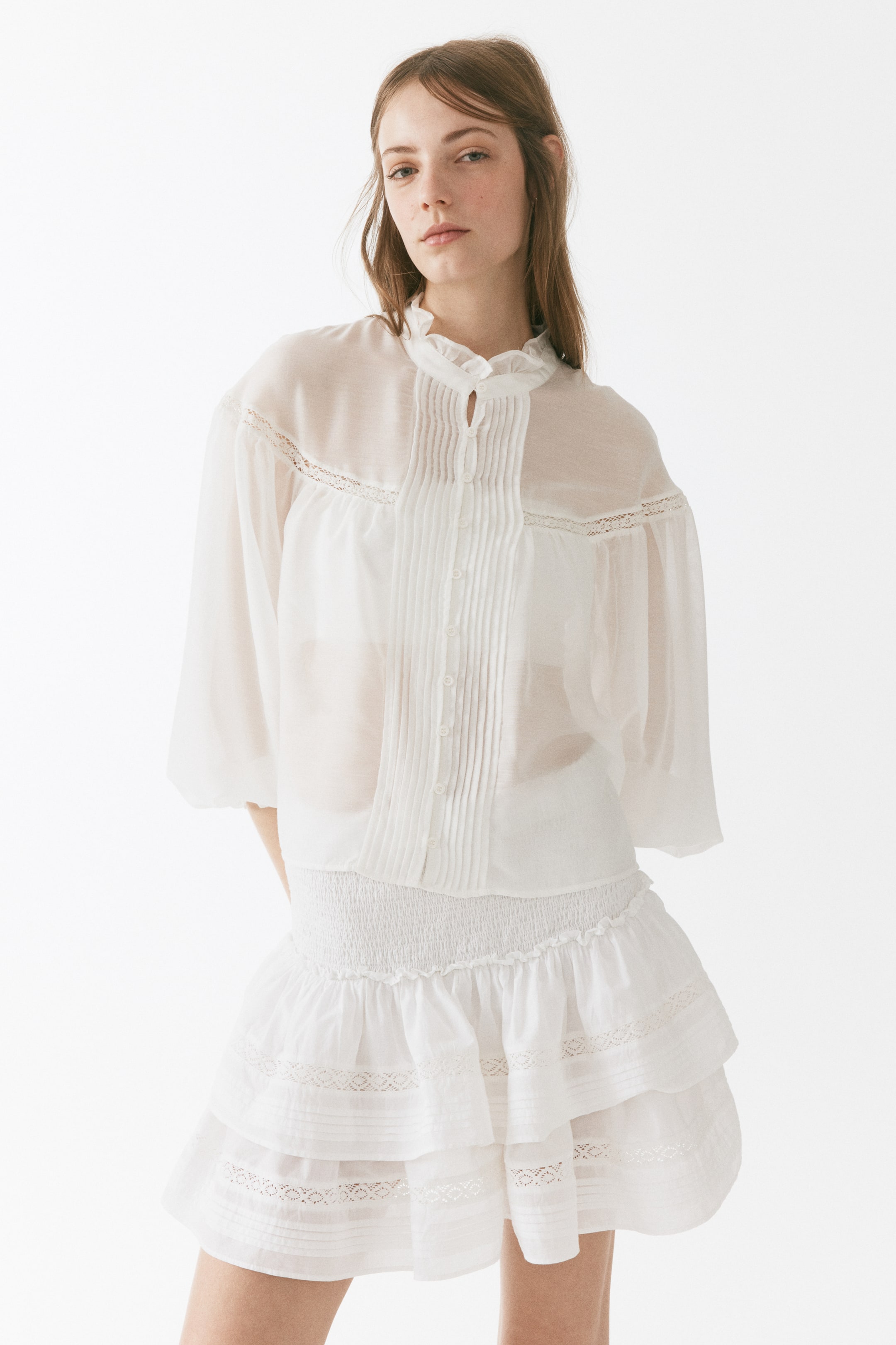View larger image: A white sheer blouse and a white tiered skirt are displayed from the front. The blouse features a ruffled stand-up collar, a button-front placket with vertical pintucks, voluminous three-quarter length sleeves, and lace detailing along the shoulder seams. The tiered skirt showcases a smocked waistband and ruffled tiers with openwork lace inserts.