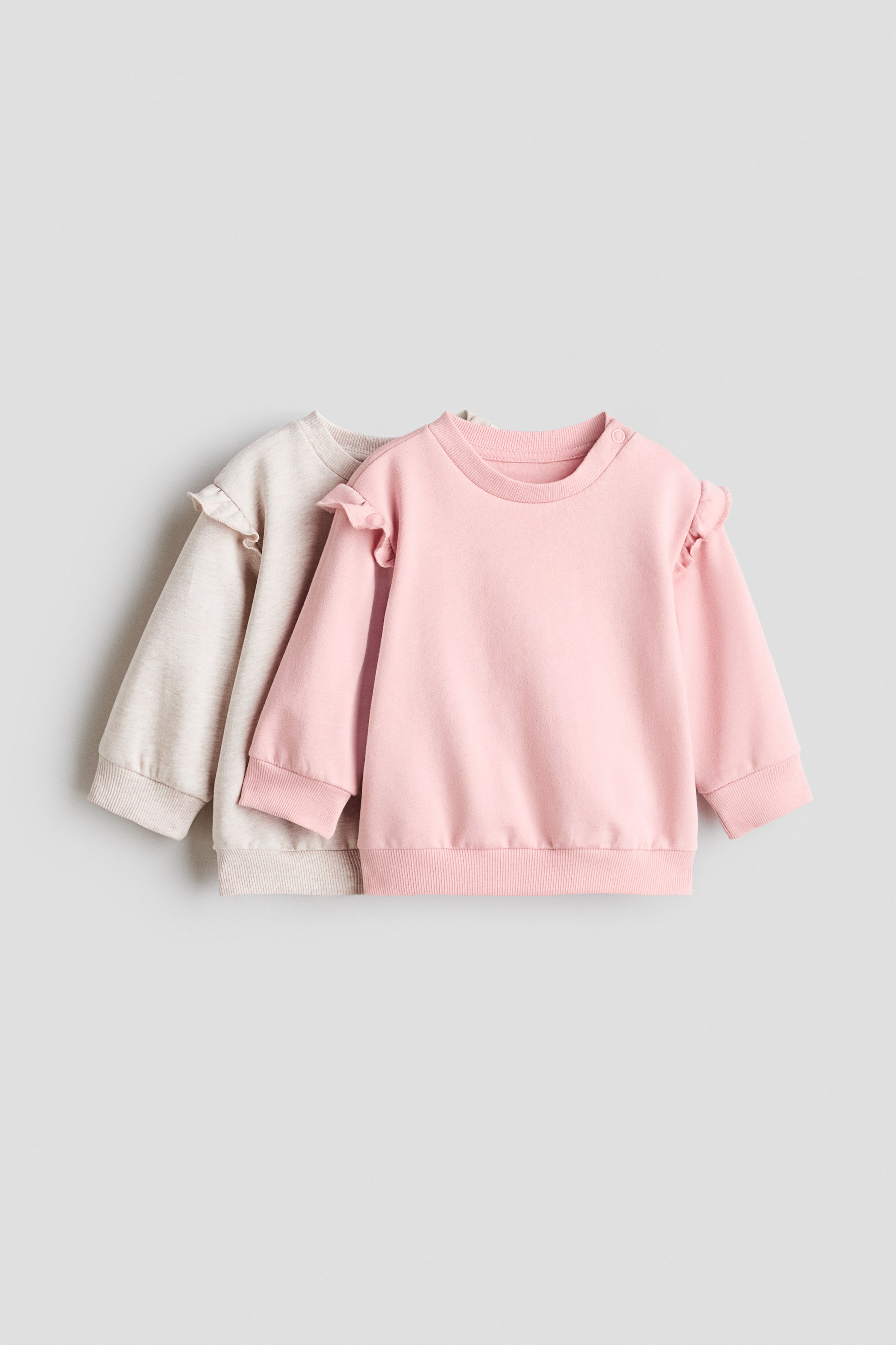 View larger image: Two kids' long-sleeved sweatshirts, one in pink and the other in light grey, are showcased from the front. Both feature a round ribbed neckline, ruffled shoulders, ribbed cuffs, and a ribbed hem. The pink sweatshirt additionally has a snap button on its left shoulder.