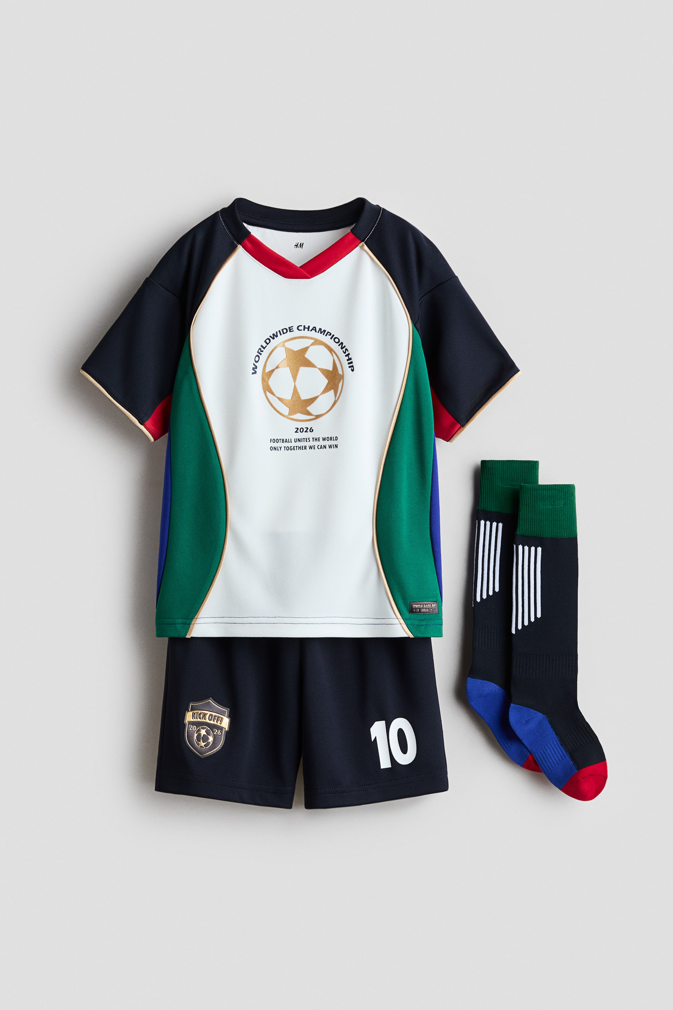 View larger image: A children's soccer uniform set is shown front-facing, featuring a multi-coloured jersey, dark blue shorts, and dark blue knee-high socks. The jersey showcases a white central panel with a gold soccer ball graphic and 'Worldwide Championship' text, bordered by gold piping and colour-blocked dark blue, red, and green panels. The shorts highlight a gold shield graphic and a white number '10', while the socks display green cuffs, white stripes, and red and blue accents.