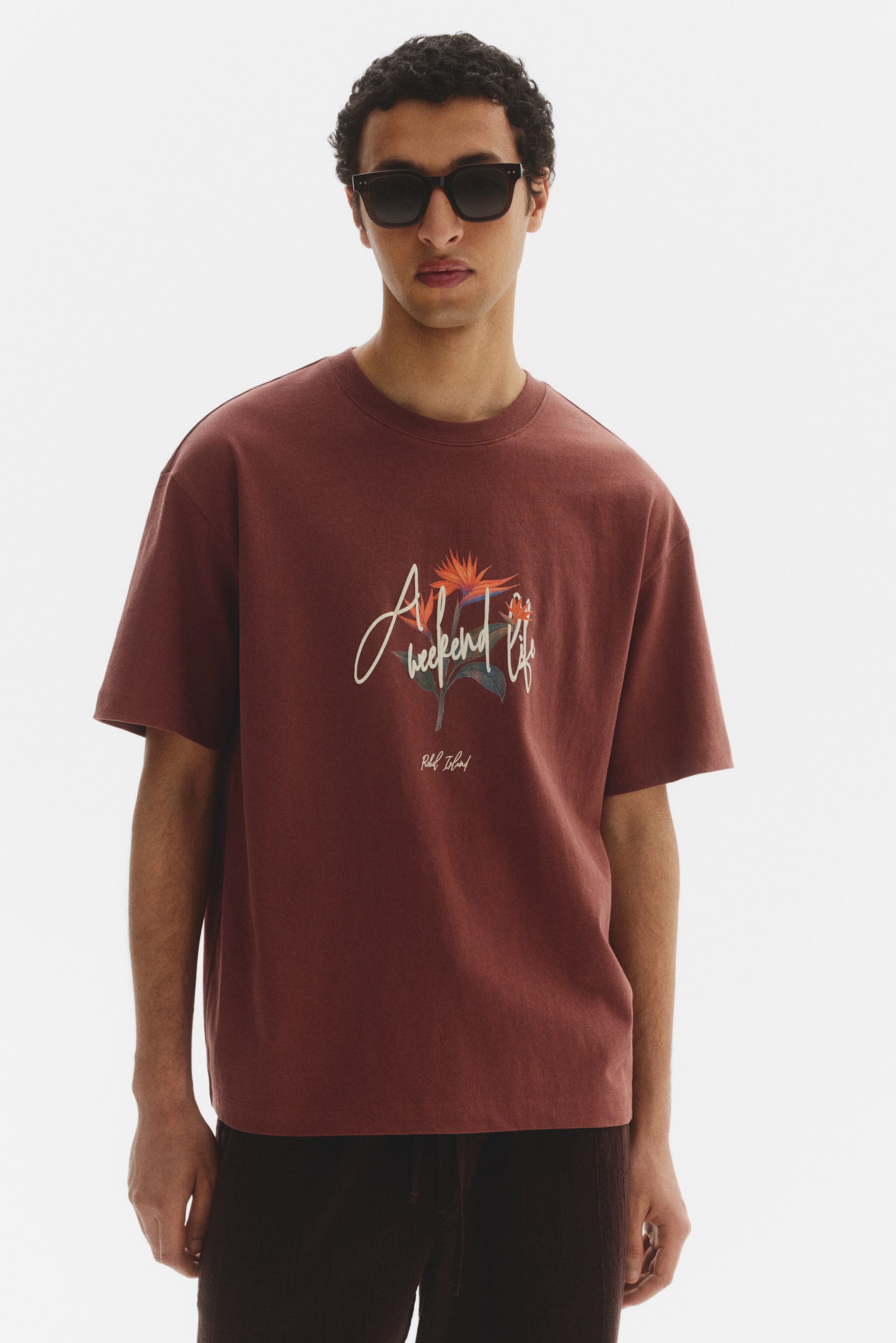 View larger image: A rust-coloured t-shirt with a relaxed fit features a crew neck and short sleeves. It showcases a central graphic with the text 'A weekend life' in script, tropical bird of paradise flowers, and 'Rebel Island' below.
