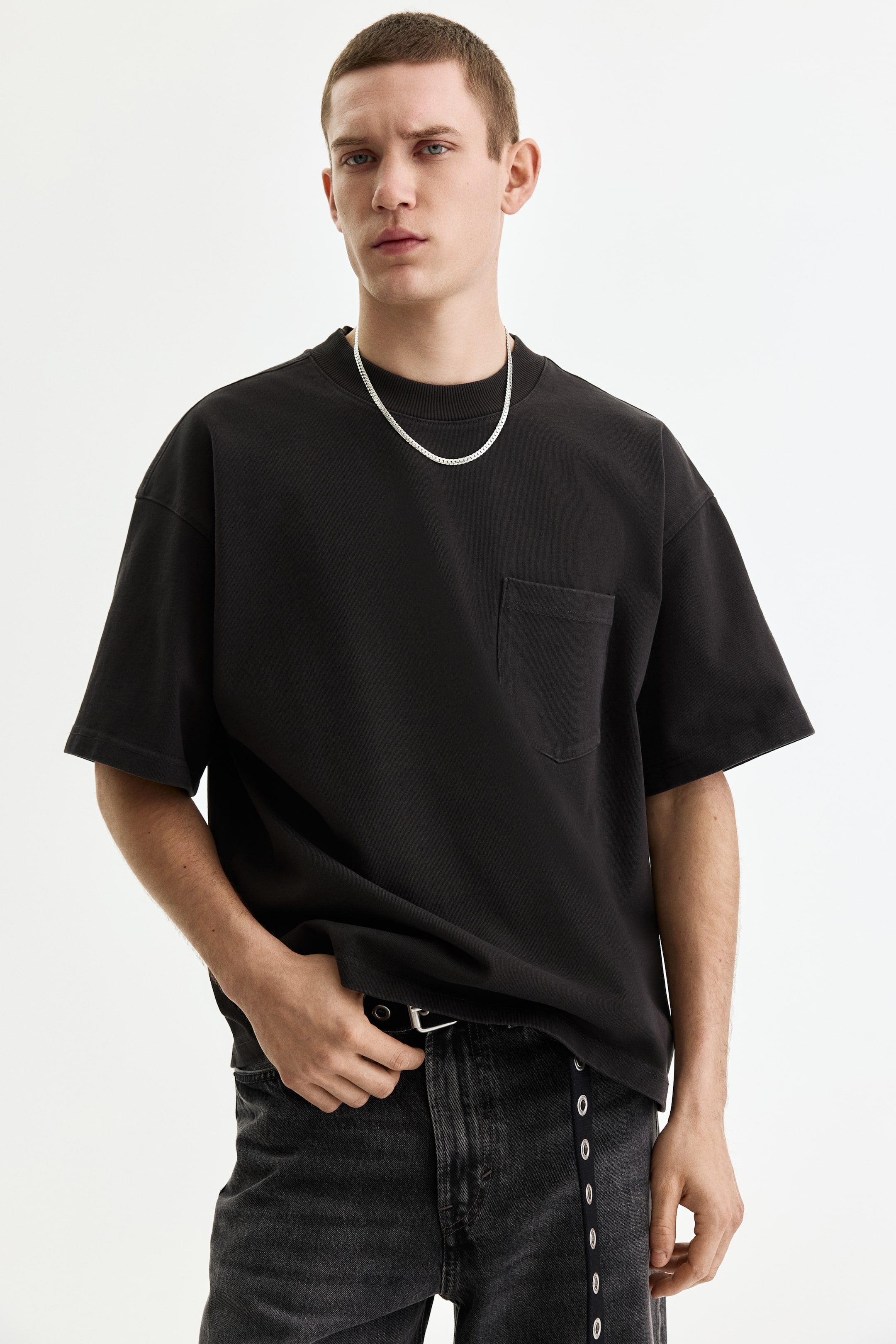 View larger image: A black oversized T-shirt is worn, featuring a ribbed crewneck and a patch pocket on the left chest. A silver chain necklace rests on the T-shirt's neckline.
