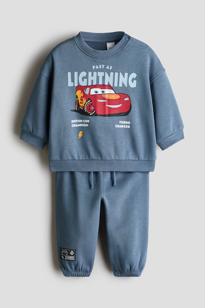 This two-piece printed sweat fabric set in dusty blue is shown from the front, featuring a long-sleeved sweatshirt and matching joggers. The sweatshirt displays a graphic print of the character Lightning McQueen on the chest alongside the text "FAST AS LIGHTNING" and is finished with a ribbed crew neck, cuffs, and hem. The coordinating joggers feature an elasticated drawstring waistband, elasticated hems, and a small black patch on the lower leg. - 1