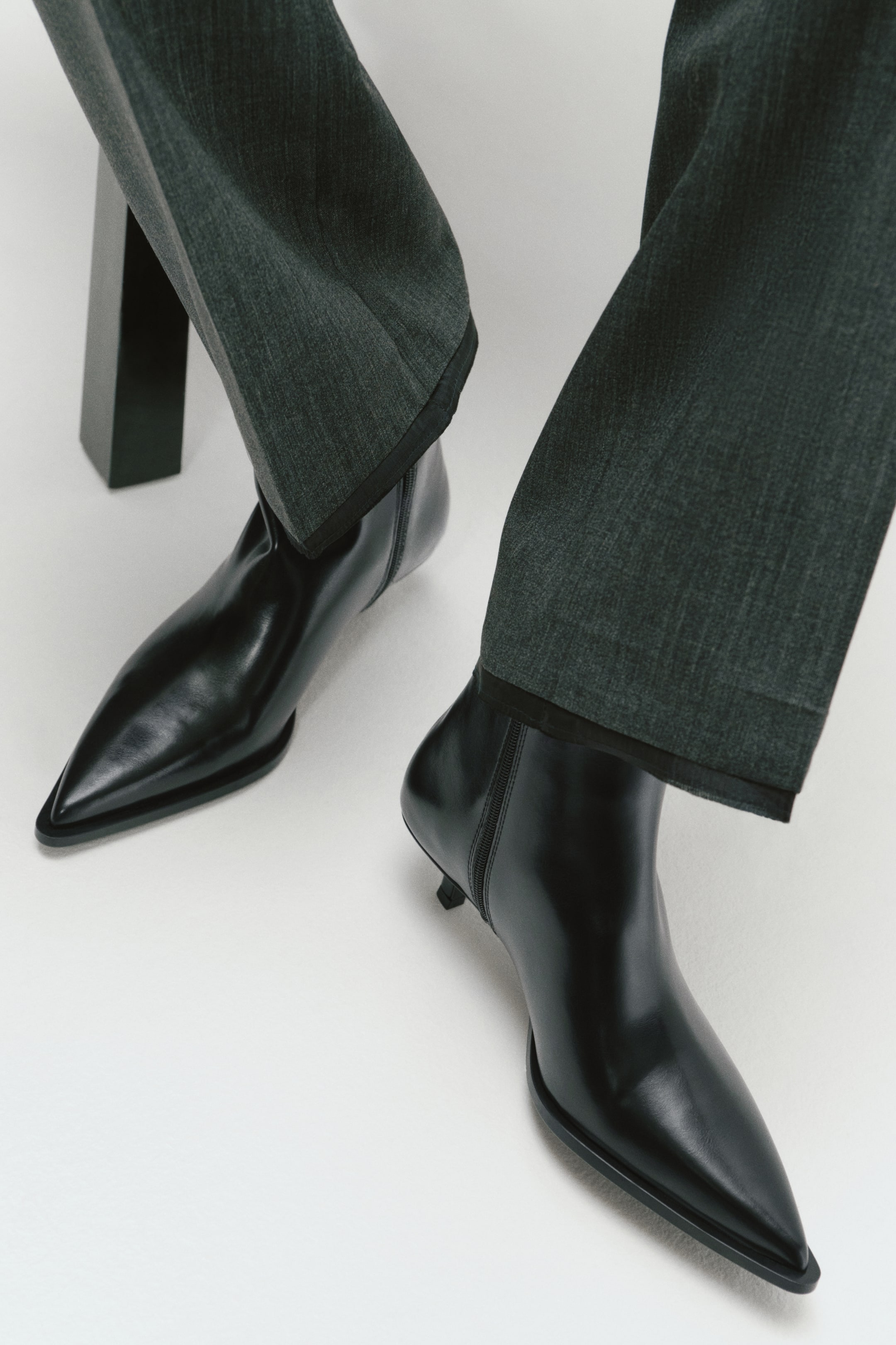 View larger image: Black leather ankle boots featuring pointed toes, low kitten heels and a side zip are being worn by the model. The boots are layered under dark grey, textured trousers.