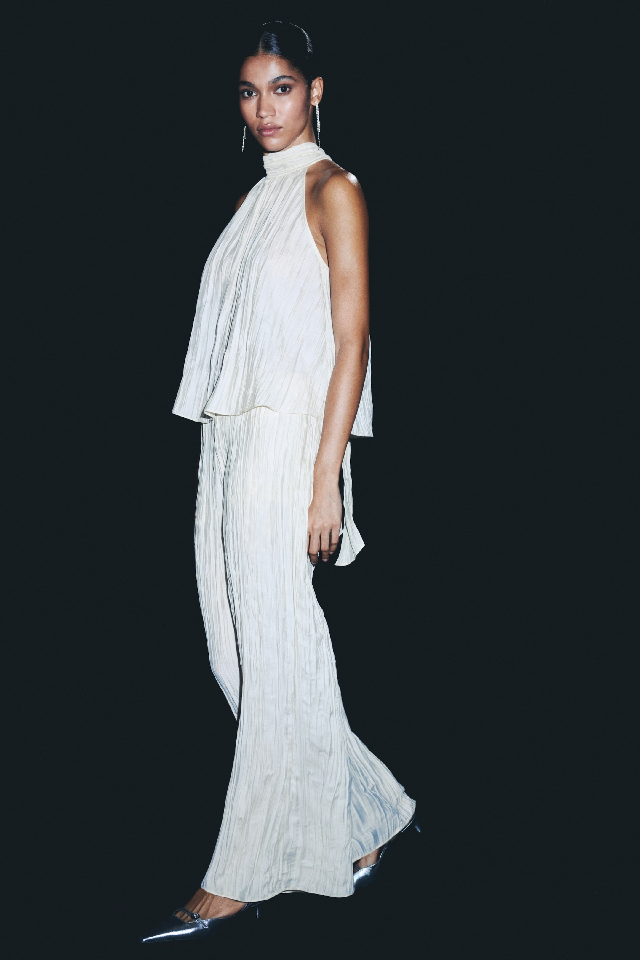 View larger image: An off-white crinkled two-piece set is being displayed from a side-front angle. The halter-neck top is rising into a high collar and is draping in a loose, layered silhouette, with a tie detail hanging from the side. The matching wide-leg pants are flowing to the floor, partially covering silver pointed-toe heels.