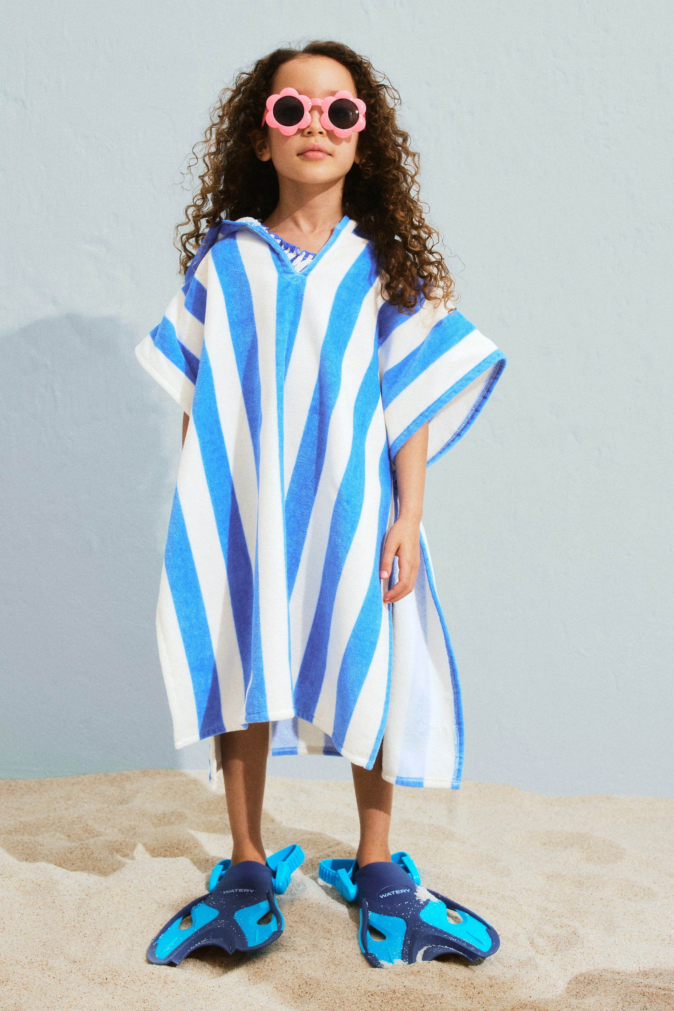 拡大画像を表示: 青と白のストライプのテリーフード付きポンチョが前向きに配置されており、幅広の縦縞、Vネックの開口部、短くて幅広の袖が特徴です。