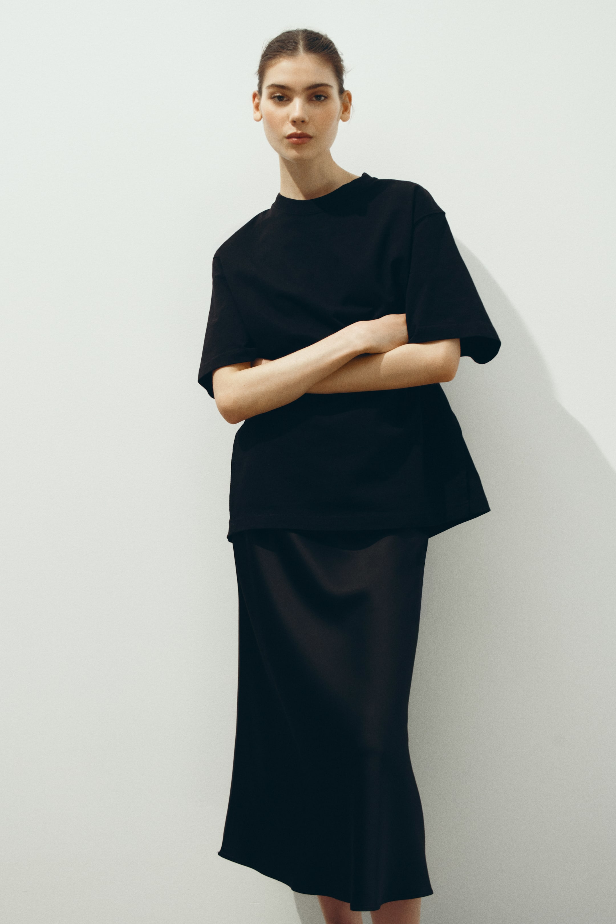 Grotere afbeelding bekijken: Een zwarte oversized T-shirt met ronde hals en brede korte mouwen wordt gepresenteerd, met een rechte zoom die de bovenkant van een zwarte midi sliprok bedekt. De rok toont een soepele val en subtiele glans, waarbij beide kledingstukken vanuit een lichte voor-rechterhoek worden gepresenteerd.