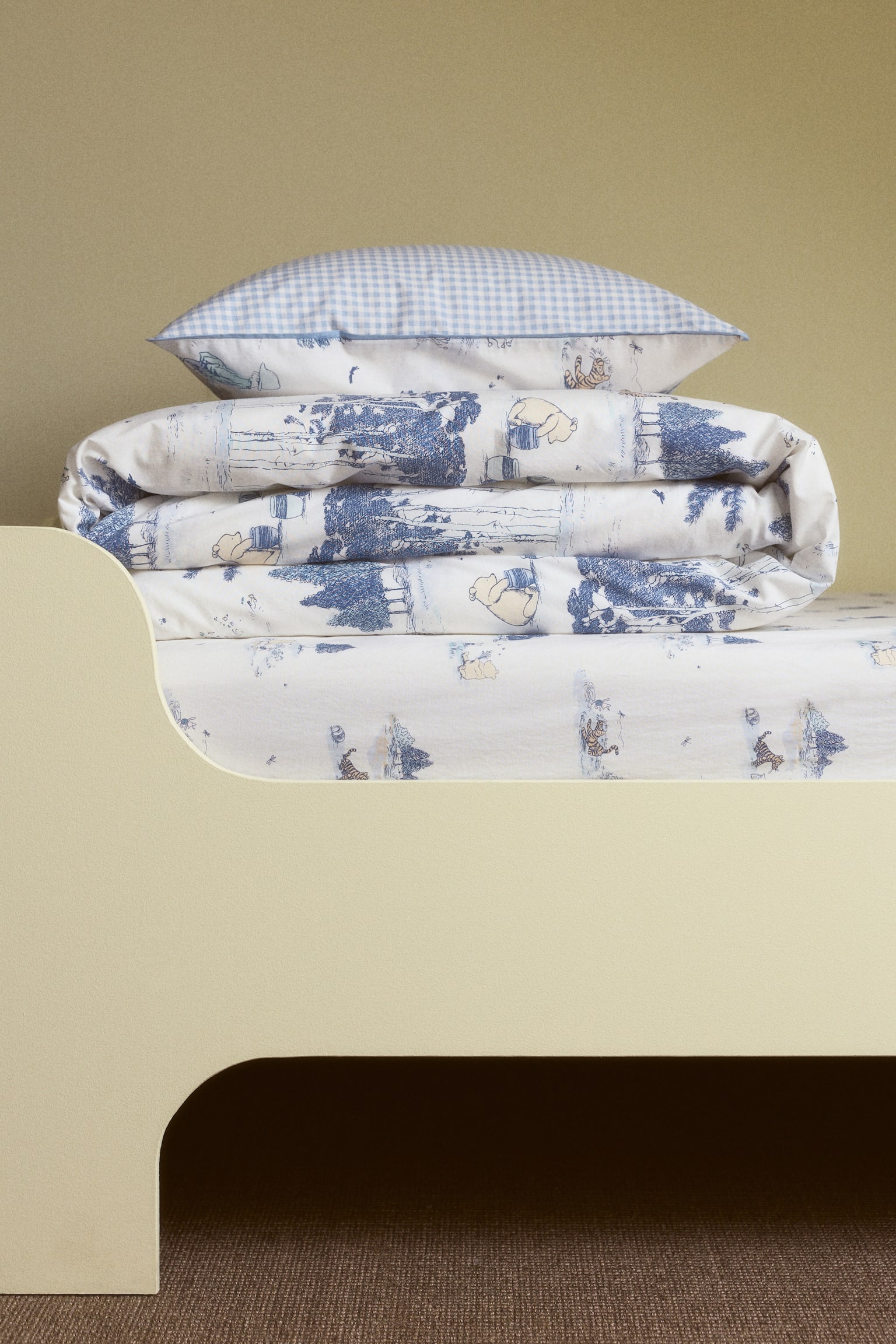 L'ensemble de literie comprend un oreiller, une couette pliée et un drap-housse, tous vus de face. L'oreiller arbore un motif vichy bleu clair et blanc sur sa face supérieure, tandis que son dessous, la couette et le drap présentent une base blanche avec un motif toile de Jouy bleu représentant des arbres et des bâtiments, entrecoupés d'illustrations de personnages de dessin animé jaunes. - 1