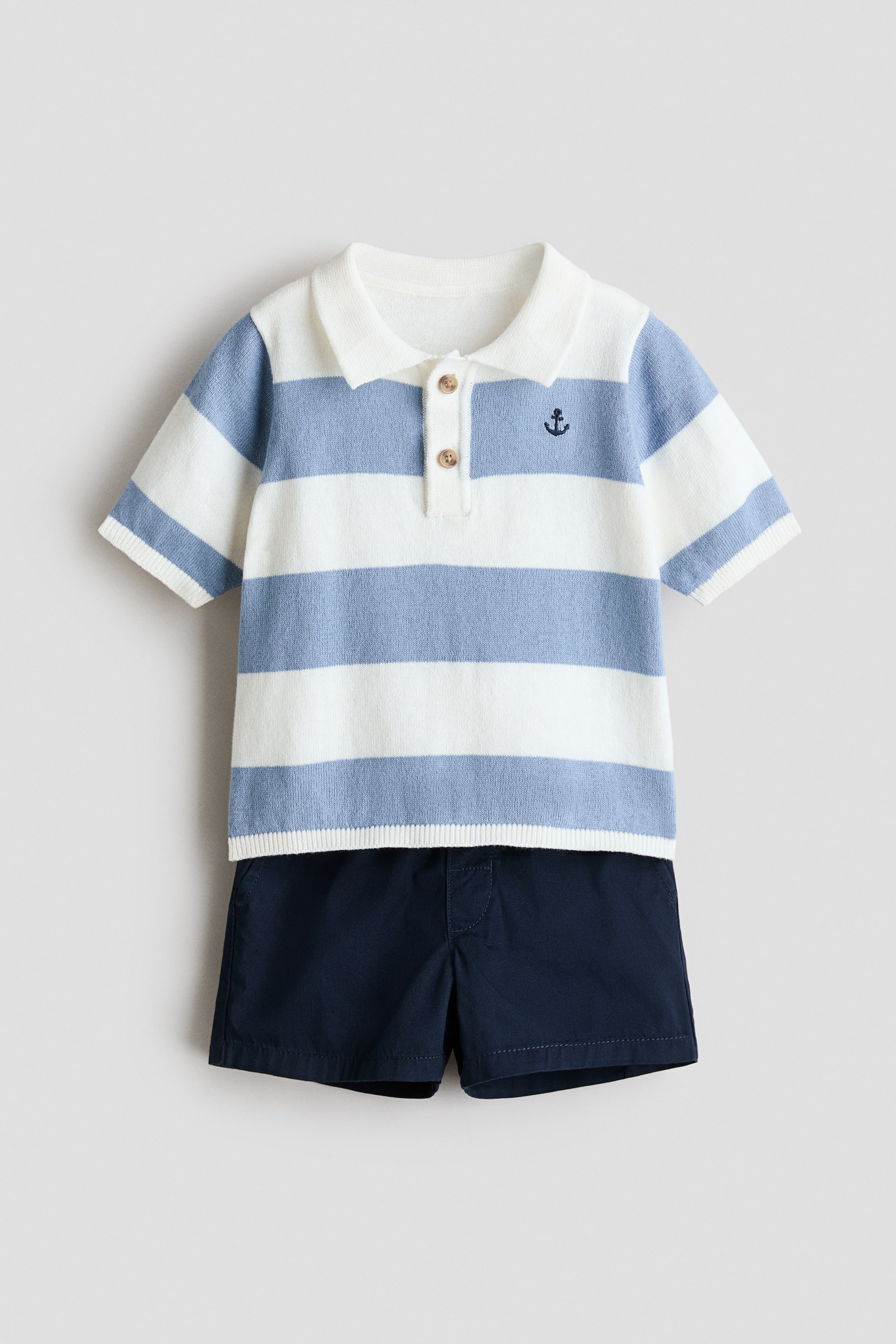 View larger image: A two-piece baby outfit is facing front, featuring a white and light blue horizontally striped knitted polo shirt with a white collar, a two-button placket with light brown buttons, a dark blue embroidered anchor on the left chest, and white ribbed cuffs and hem. This polo shirt is layered over dark blue shorts, which are displaying an elasticated waistband.