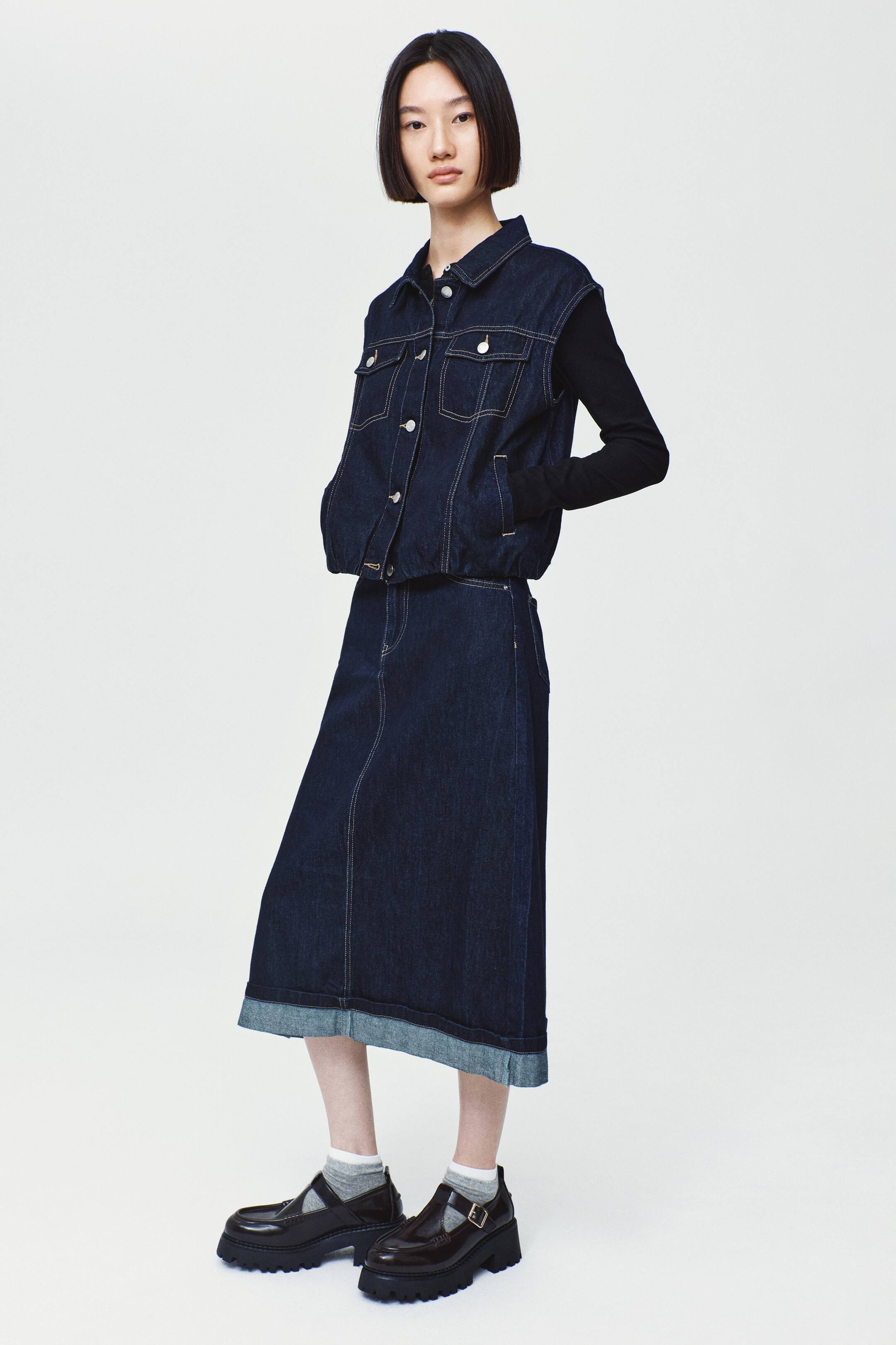 View larger image: The model is wearing a dark blue, calf-length denim skirt with a visible seam running down the front and a wide, contrasting turn-up hem. The skirt is styled with a matching dark denim vest layered over a black long-sleeve shirt.