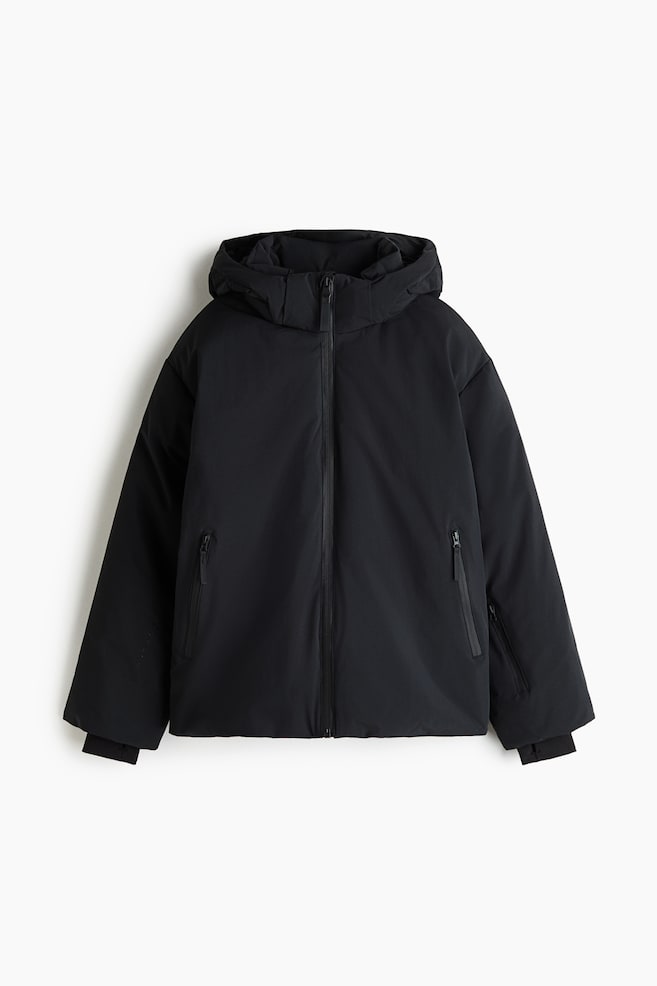 A black hooded jacket is shown from the front, featuring a smooth matte shell, a full-length centre zip, and a high-collar hood. The jacket features vertical zipped hand pockets on each side and ribbed storm cuffs extending from the sleeve hems. - 1