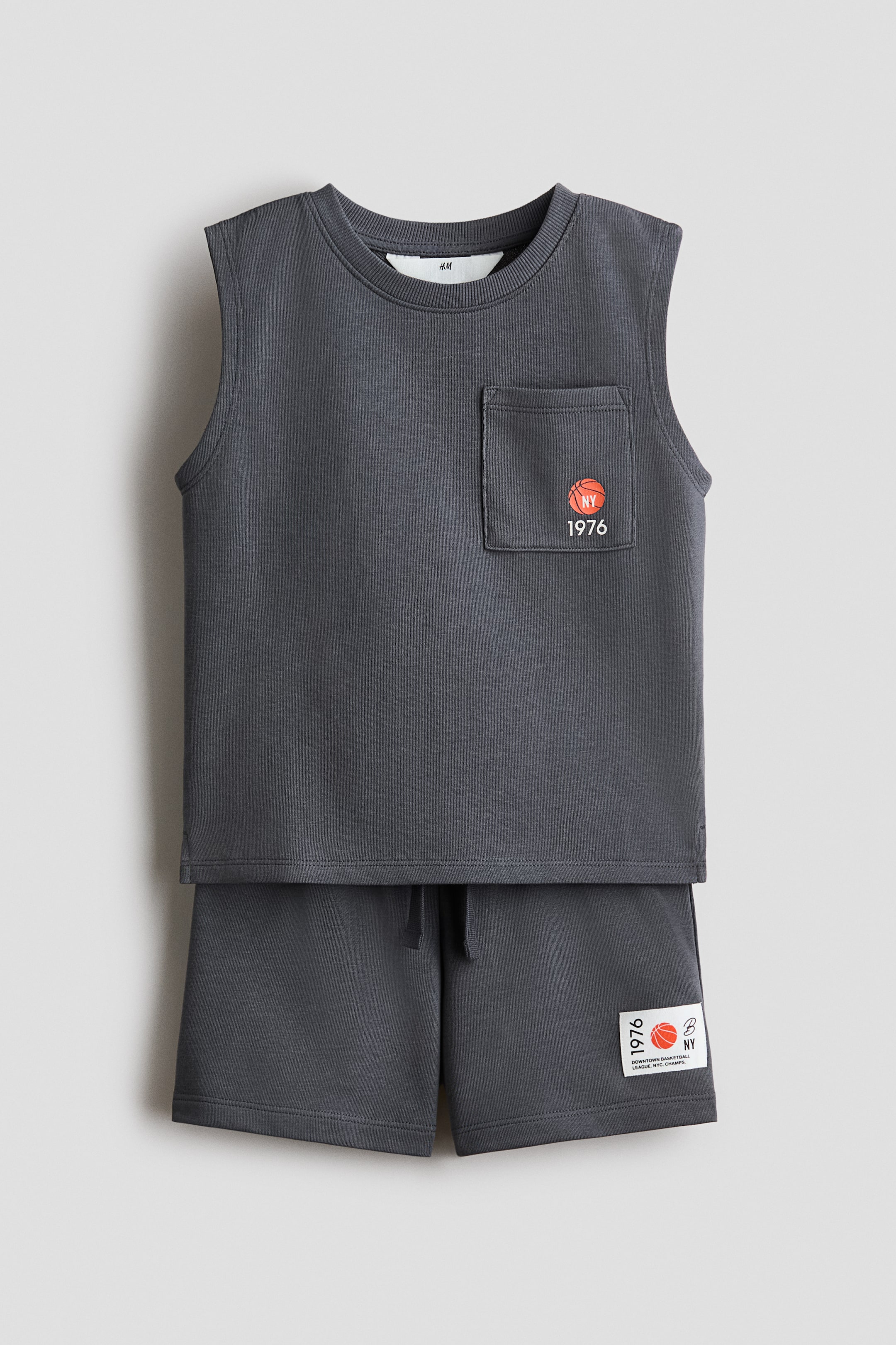 View larger image: Introducing a dark gray two-piece set, featuring a sleeveless top with a ribbed crew neck and a chest patch pocket showcasing an orange basketball graphic with the text 'NY 1976'. The matching shorts come with a drawstring waist and a rectangular patch on the lower left leg, displaying '1976', an orange basketball graphic, and 'NY' text.