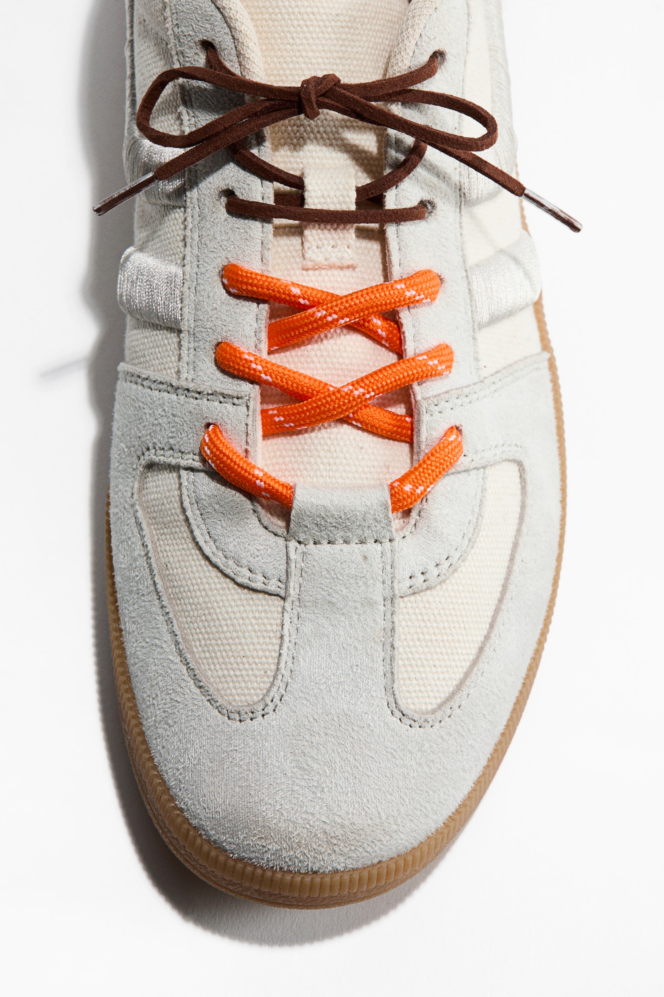 Grotere afbeelding bekijken: Aan de voorkant van een sneaker worden twee paar veters getoond die verschillende stijlen demonstreren. Een paar platte, donkerbruine veters is bovenaan in een strik geknoopt, terwijl een paar ronde, feloranje veters met witte spikkels kruislings door de onderste vetergaatjes loopt.