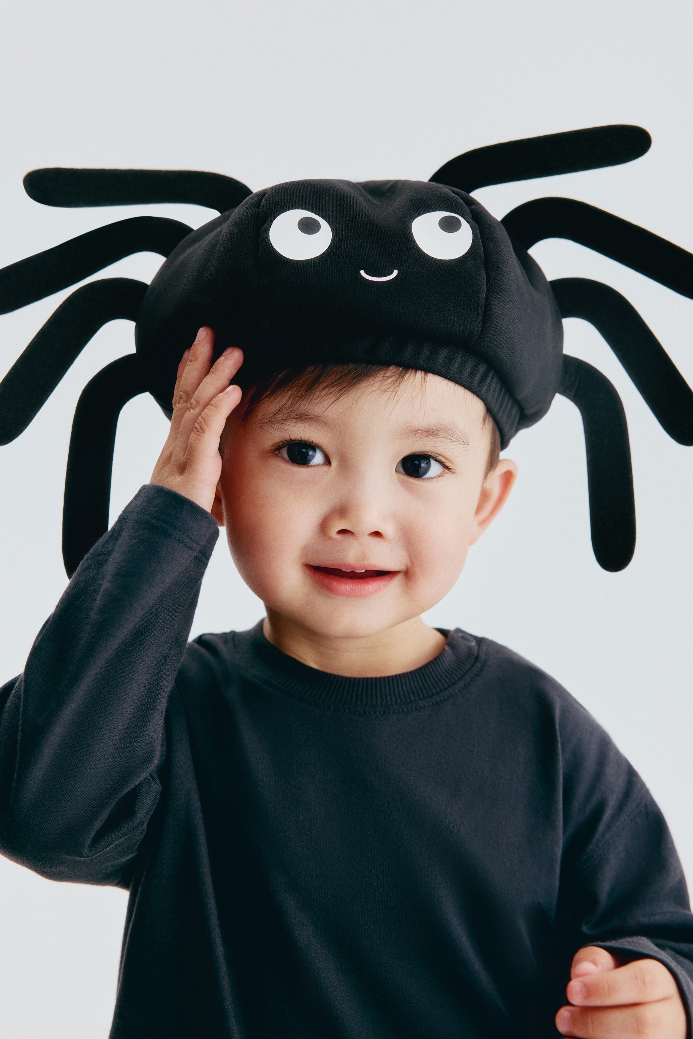 Un sombrero negro acolchado de Halloween está diseñado como una araña, mirando hacia el frente. Está mostrando un cuerpo redondeado con dos grandes ojos blancos con pupilas negras, una pequeña boca rosada sonriente y ocho patas acolchadas y curvadas que se extienden desde sus lados.