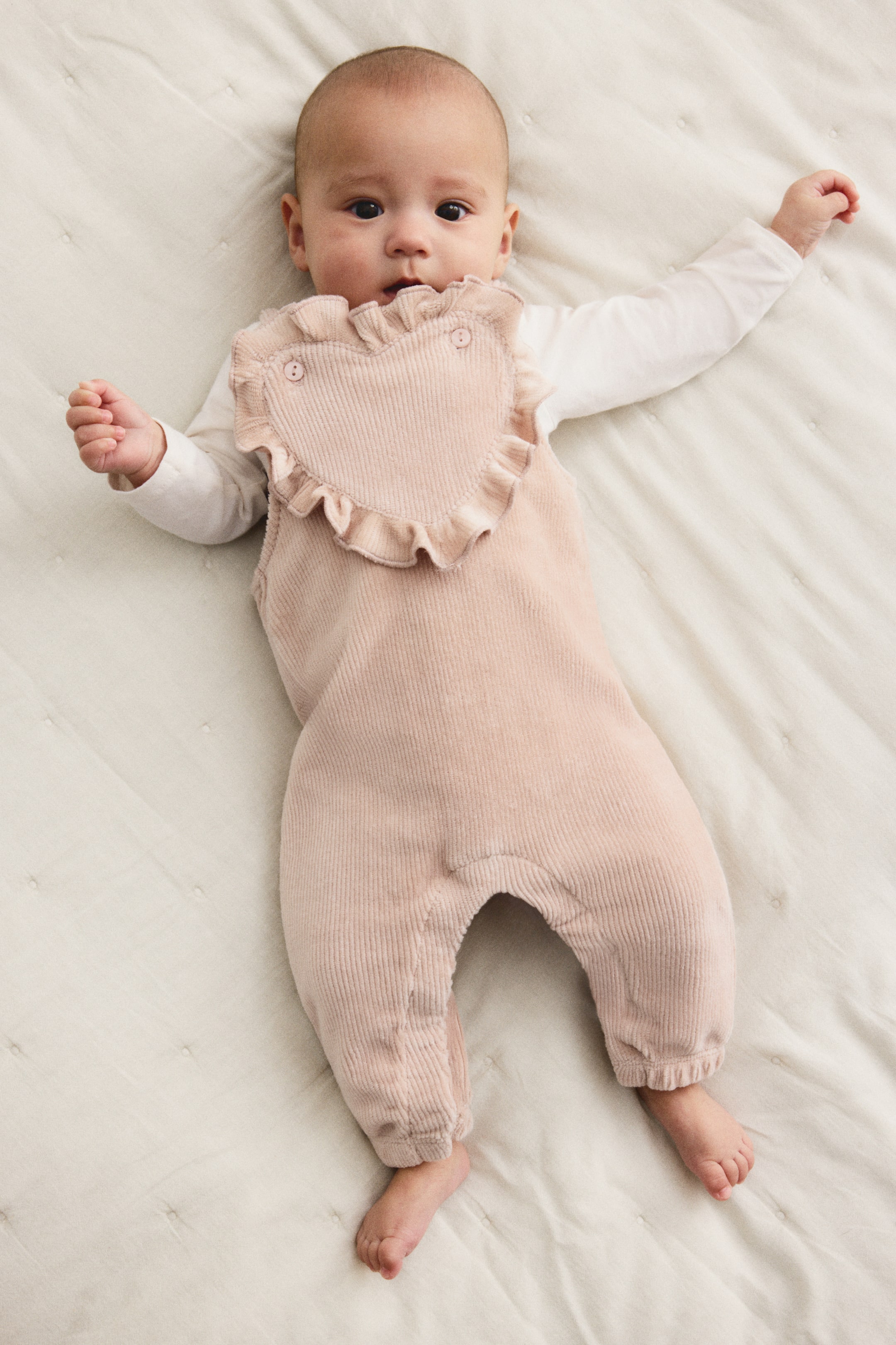 View larger image: A pink corduroy baby overall is lying flat, featuring a heart-shaped bib with ruffles and two visible buttons. It is layering over a white long-sleeve top, and the overall's full-length legs are gathering at the ankles.