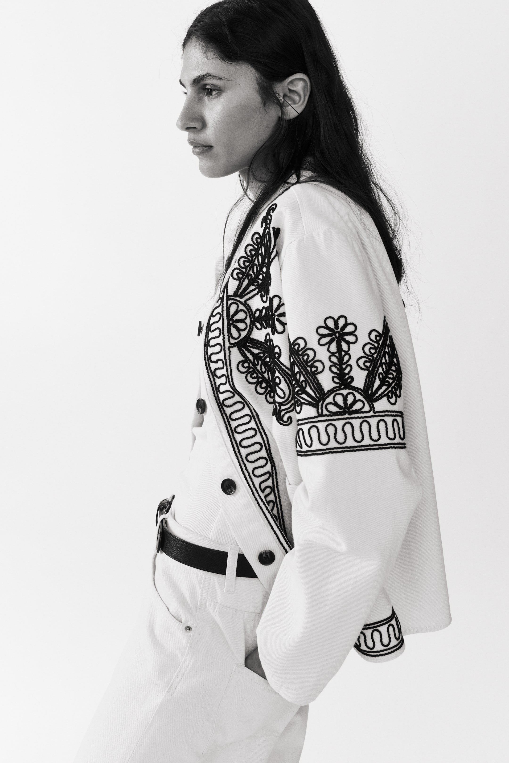 Agrandir l'image: Une veste en sergé de coton blanc avec des motifs brodés noirs est portée par le mannequin. La veste est dotée de boutons noirs sur le devant, et le mannequin place une main dans une poche avant ouverte.
