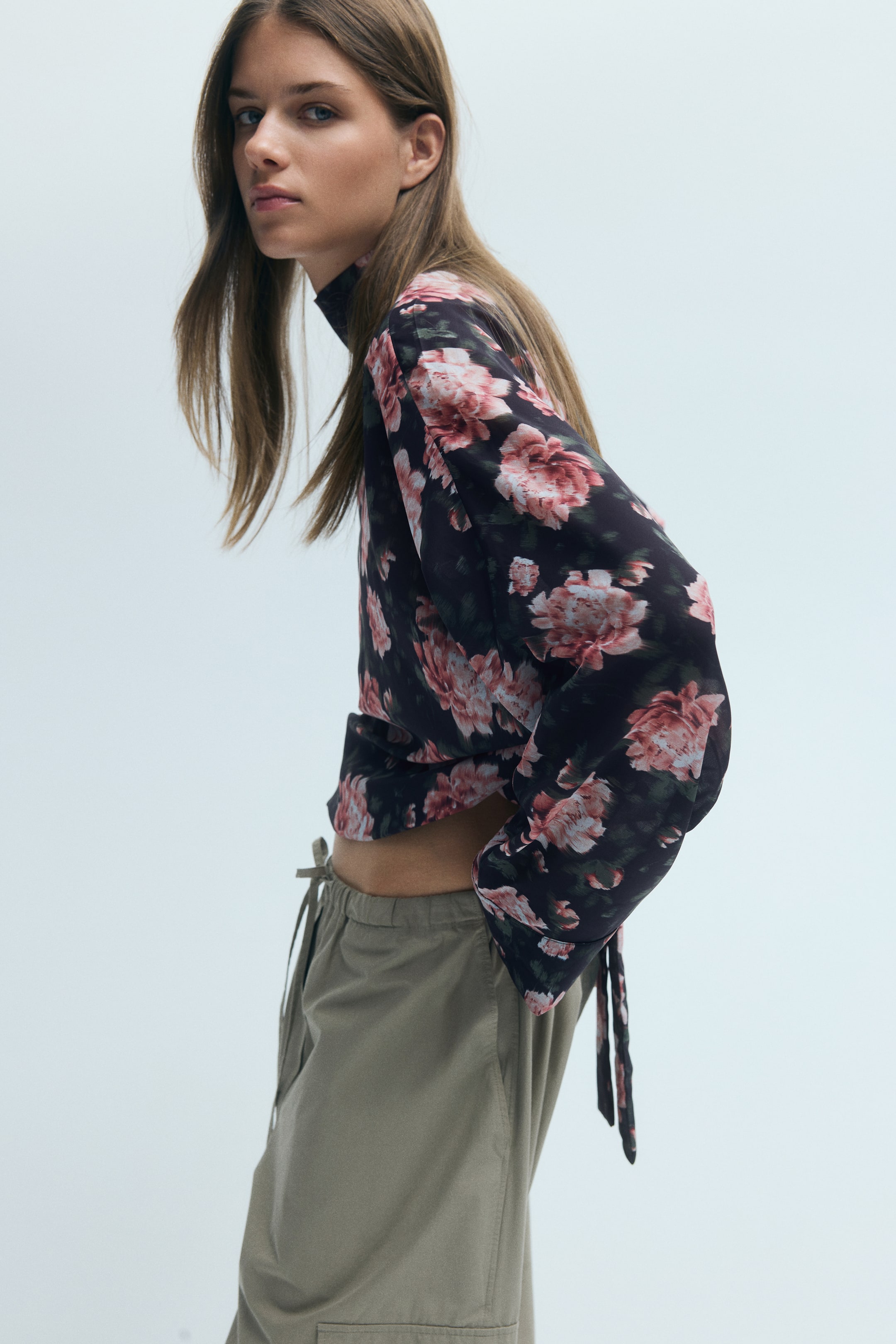 Grotere afbeelding bekijken: De model draagt een blouse met bloemenprint in een donkere, transparante weving, met een hoge bandkraag en linten die langs de achterkant naar beneden hangen. De blouse heeft verlaagde schouders, lange mouwen met manchetten en een elastische zoom, gestyled met olijfgroene trekkoordbroeken.