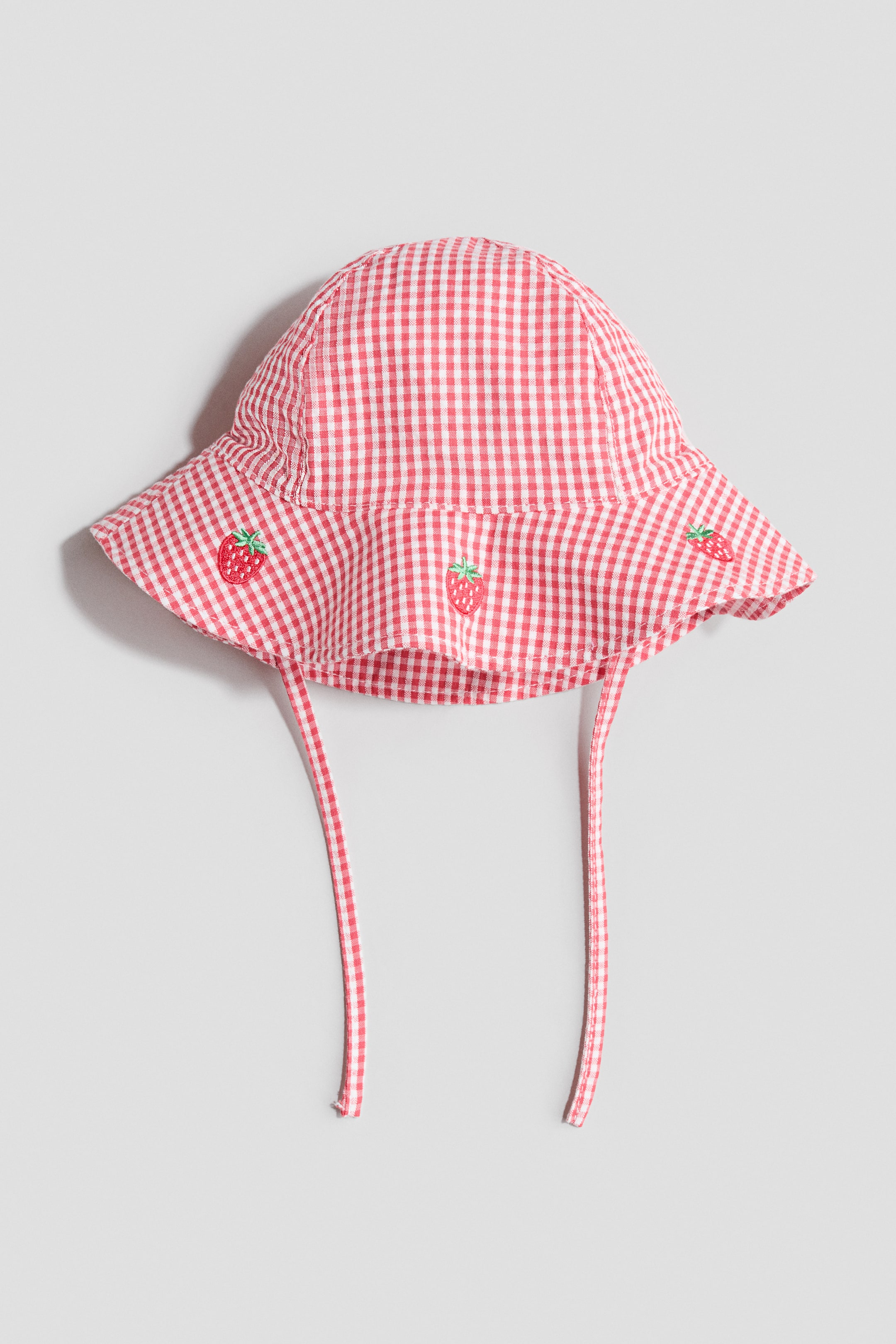 Grotere afbeelding bekijken: Een zonnehoed in zachte katoenen mousseline met een rood-wit geruite patroon. De hoed is ontworpen met een brede, zachte rand die aan de voorkant is versierd met drie geborduurde aardbeimotieven, en bijpassende geruite kinbandjes die aan beide zijden uitsteken.