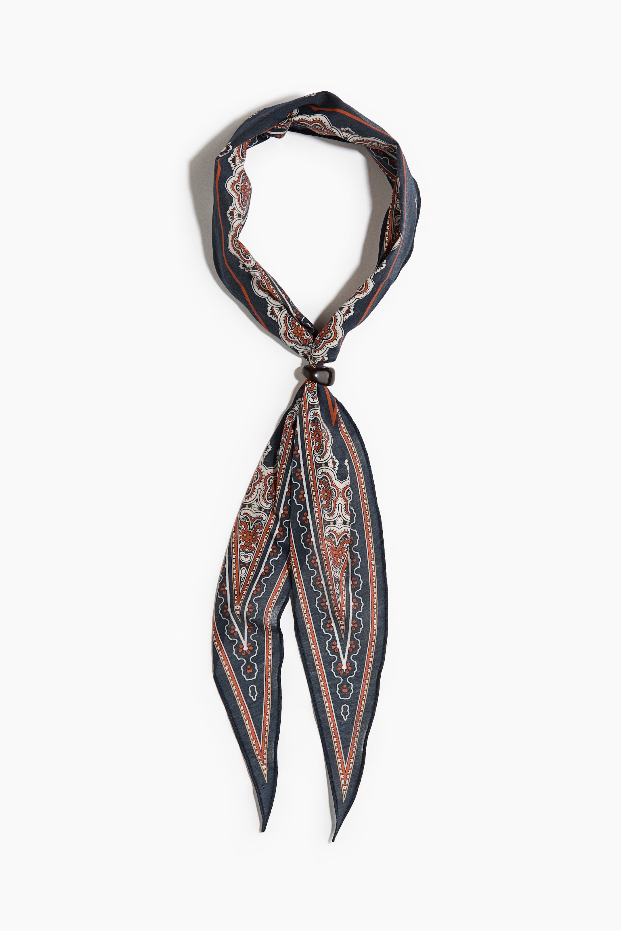View larger image: A diamond-shaped neck scarf in an airy cotton weave is displaying a dark blue background with a repeating paisley-like pattern in white and rust orange. A dark cubic metal bead is cinching the scarf, forming a loop at the top and two pointed ends hanging below.