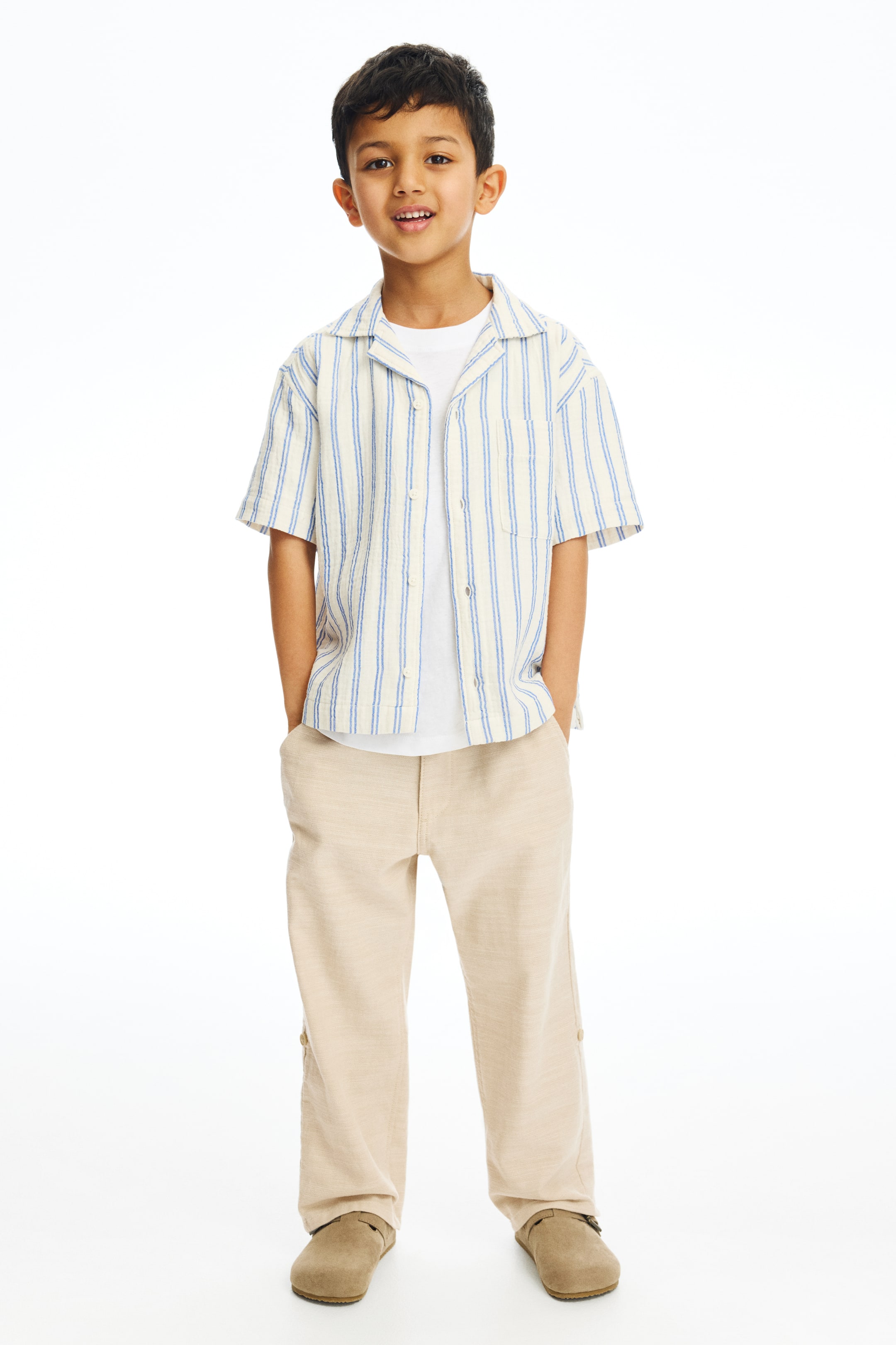 View larger image: A child's outfit consists of a short-sleeve, revere-collared shirt with vertical blue and cream stripes, worn open over a white t-shirt. Relaxed-fit light beige pants, featuring small buttons on the outer lower legs, are paired with light brown, closed-toe slip-on shoes.