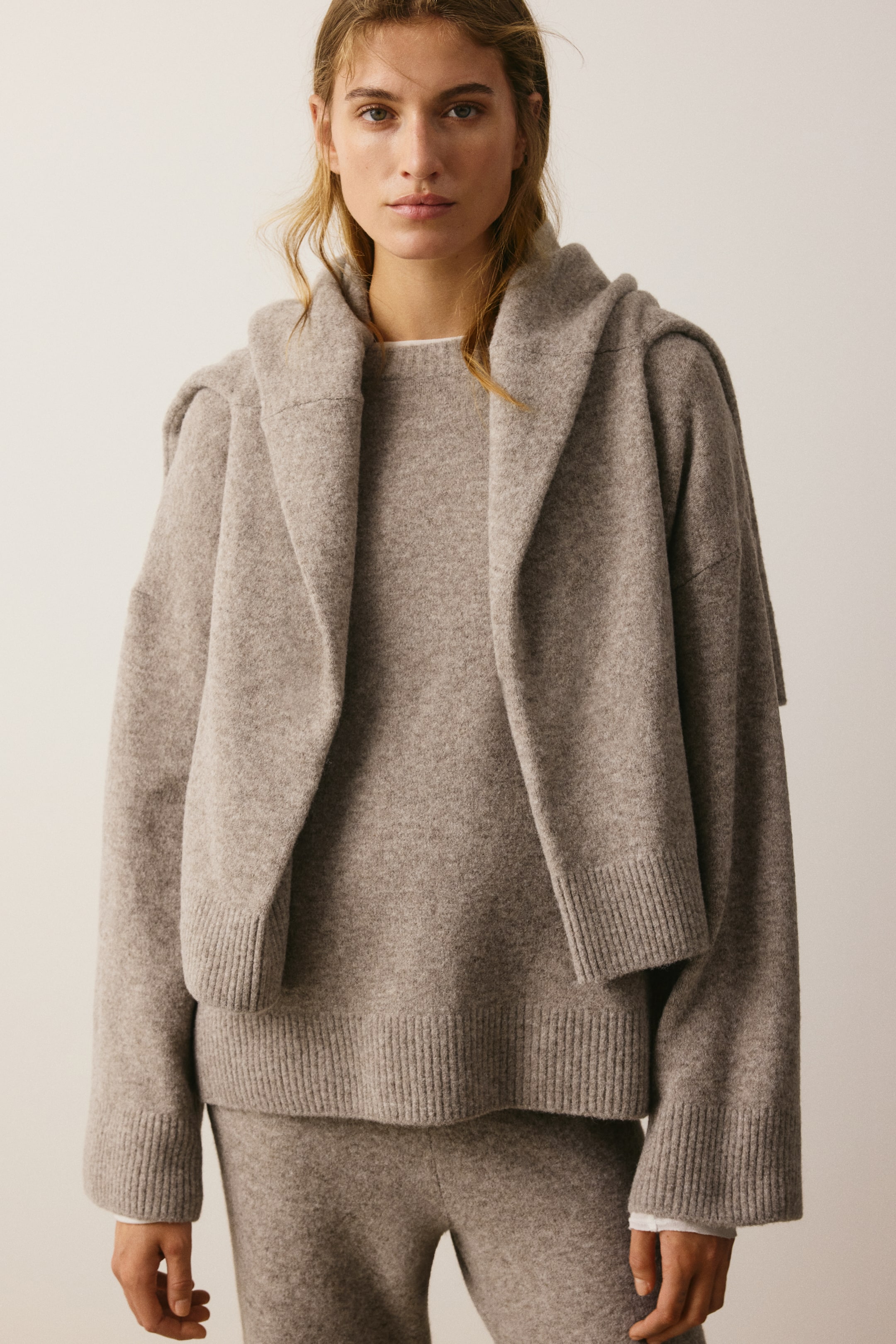 View larger image: A light-brown knit sweater is shown from the front, featuring a round ribbed neckline, dropped shoulders, and wide ribbed cuffs and hem. The relaxed-fit sweater is styled with a matching knit sweater draped over the shoulders and matching knit trousers.