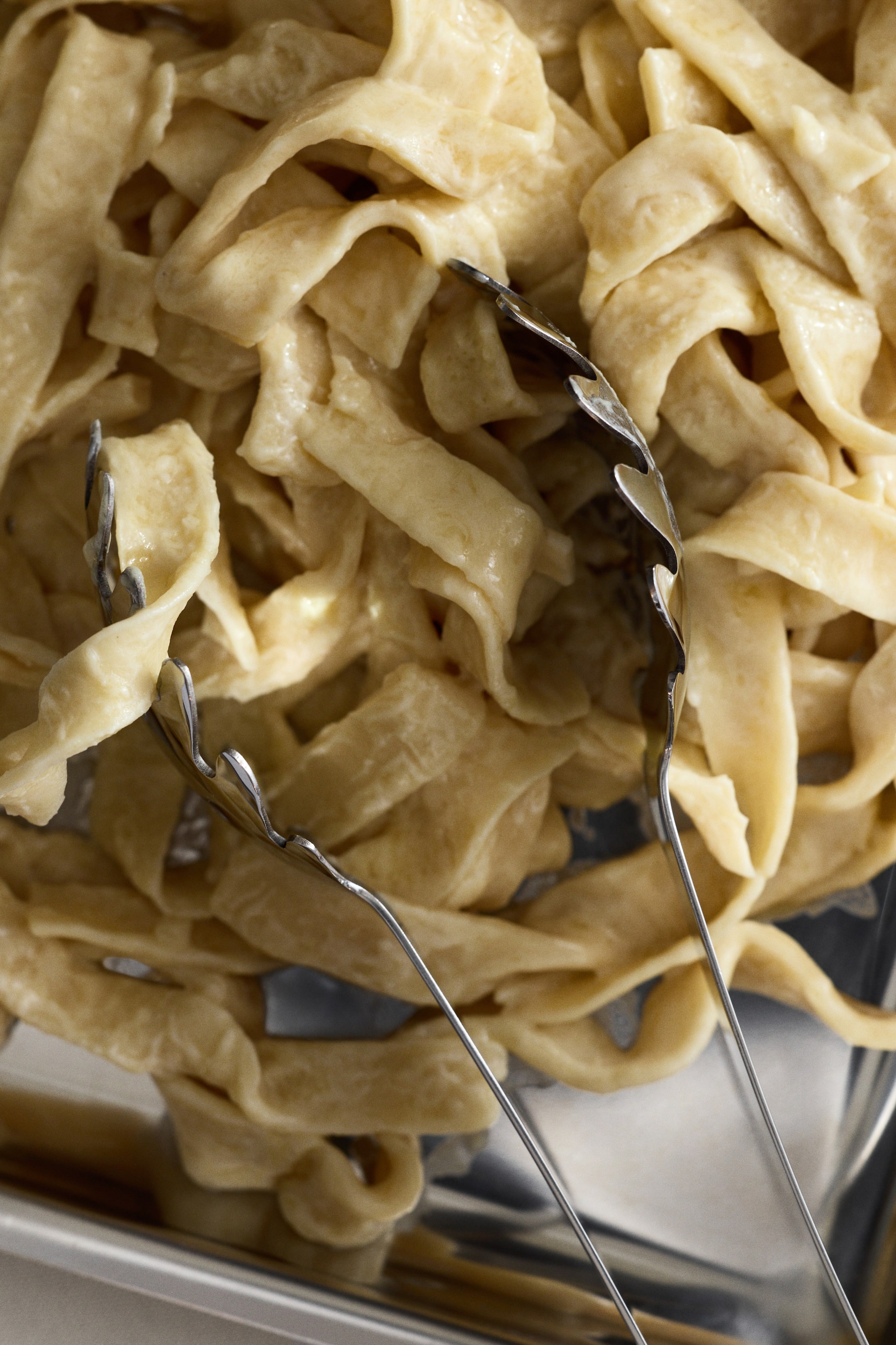 הצגת תמונה גדולה יותר: מלקחי פסטה מפלדת אל-חלד מציגים עיצוב חלק ואלגנטי, כששתי זרועותיהם מסתיימות במשטחי תפיסה פתוחים וגליים. המלקחים ממוקמים בתוך ערימה של פסטה ארוכה, ונראים כאילו הם אוחזים או עומדים לאחוז בחוטי הפסטה.