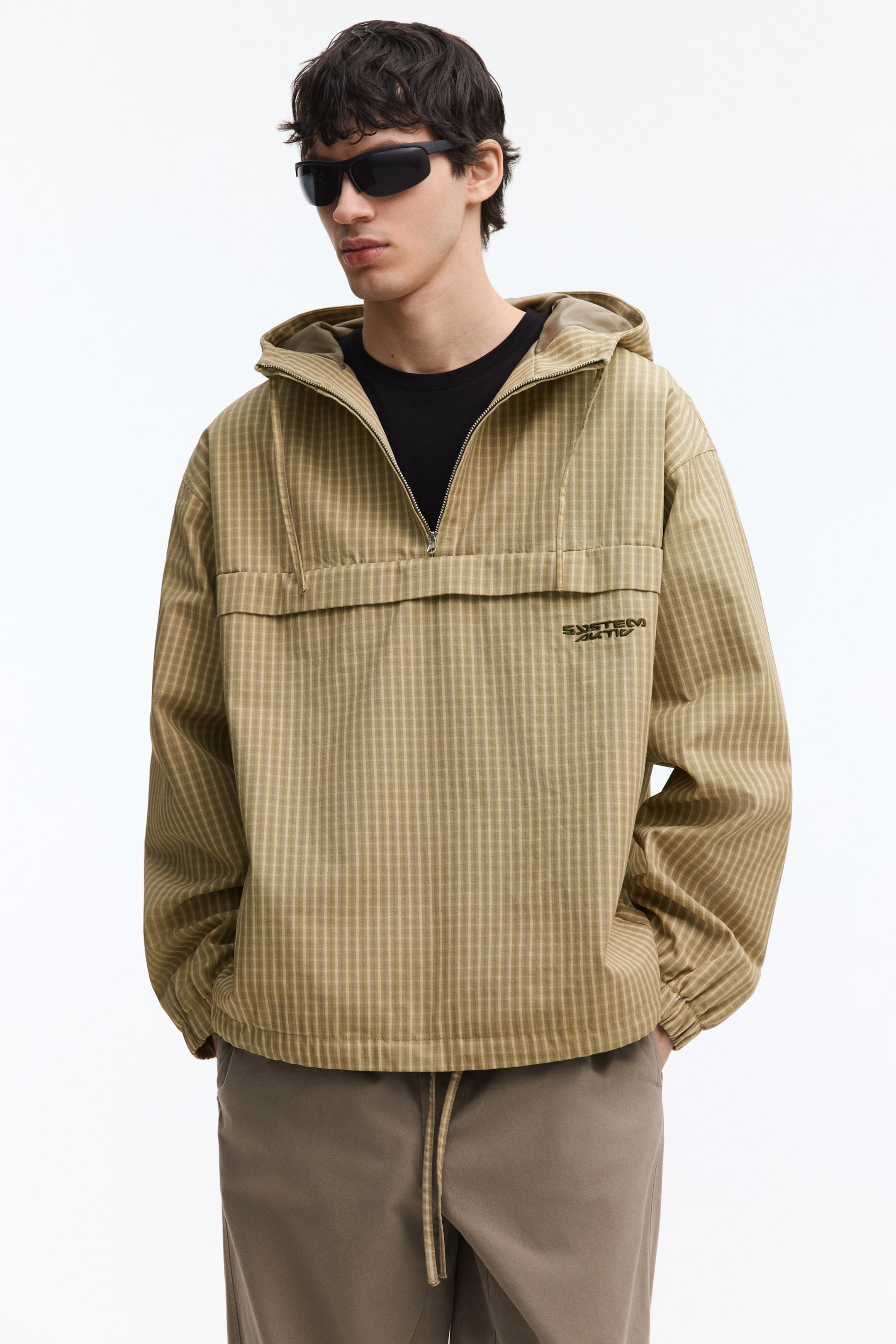 Visa större bild: En khaki-rutigt mönstrad anorak med huva presenteras från en frontvy, med en halv dragkedja, ett horisontellt klaffdetalj över bröstet och en broderad 'SYSTEM ACTIVE'-logotyp på vänster bröst. Anoraken har en avslappnad passform med elastiska muddar och fåll, och en svart t-shirt syns vid halsöppningen.