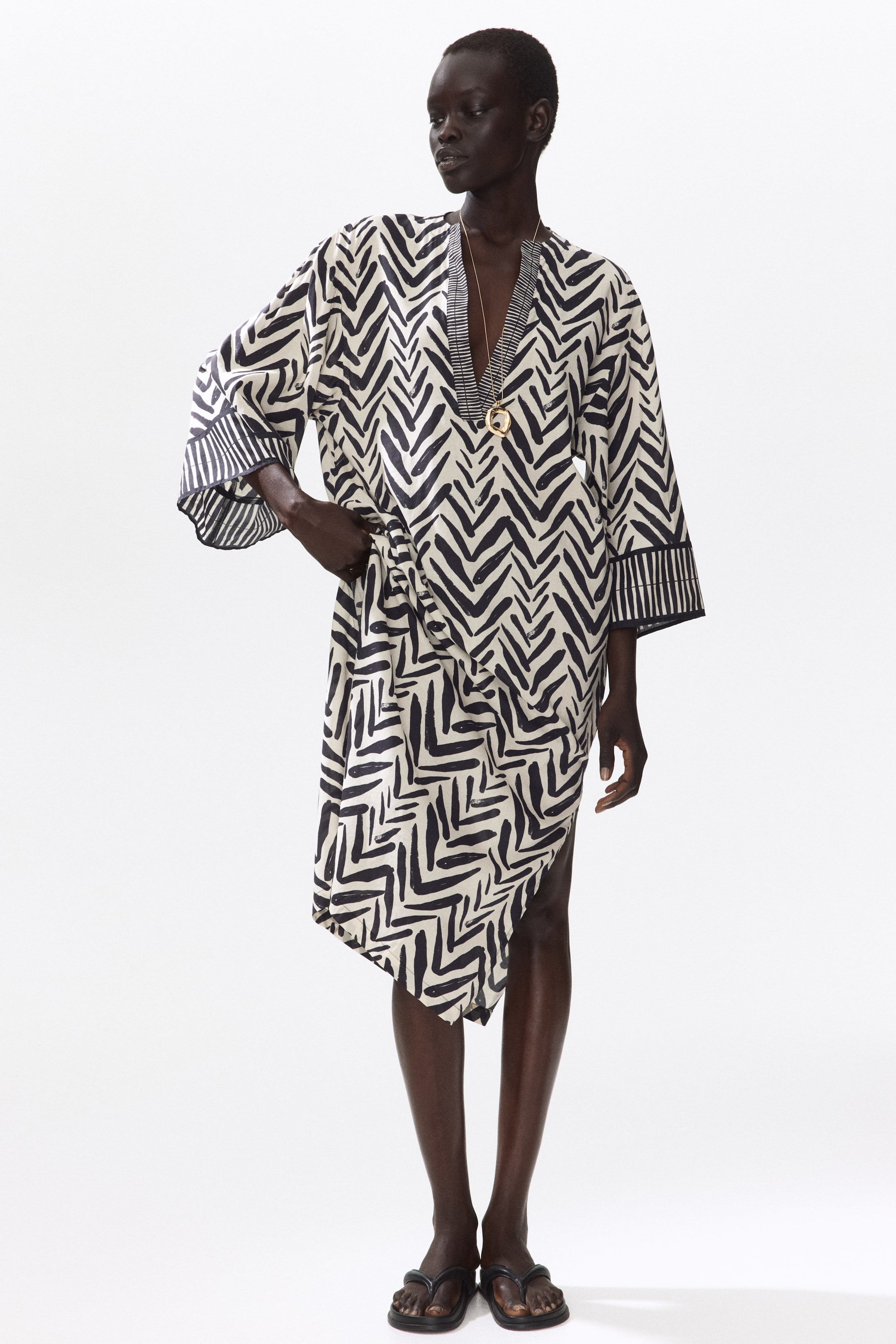 View larger image: The model is wearing a calf-length tunic dress featuring a black and off-white abstract chevron pattern, a V-neckline, and 3/4-length sleeves. The dress showcases an asymmetrical hemline with a high side slit. A long gold necklace accentuates the V-neckline, while the model's hand rests on her hip.