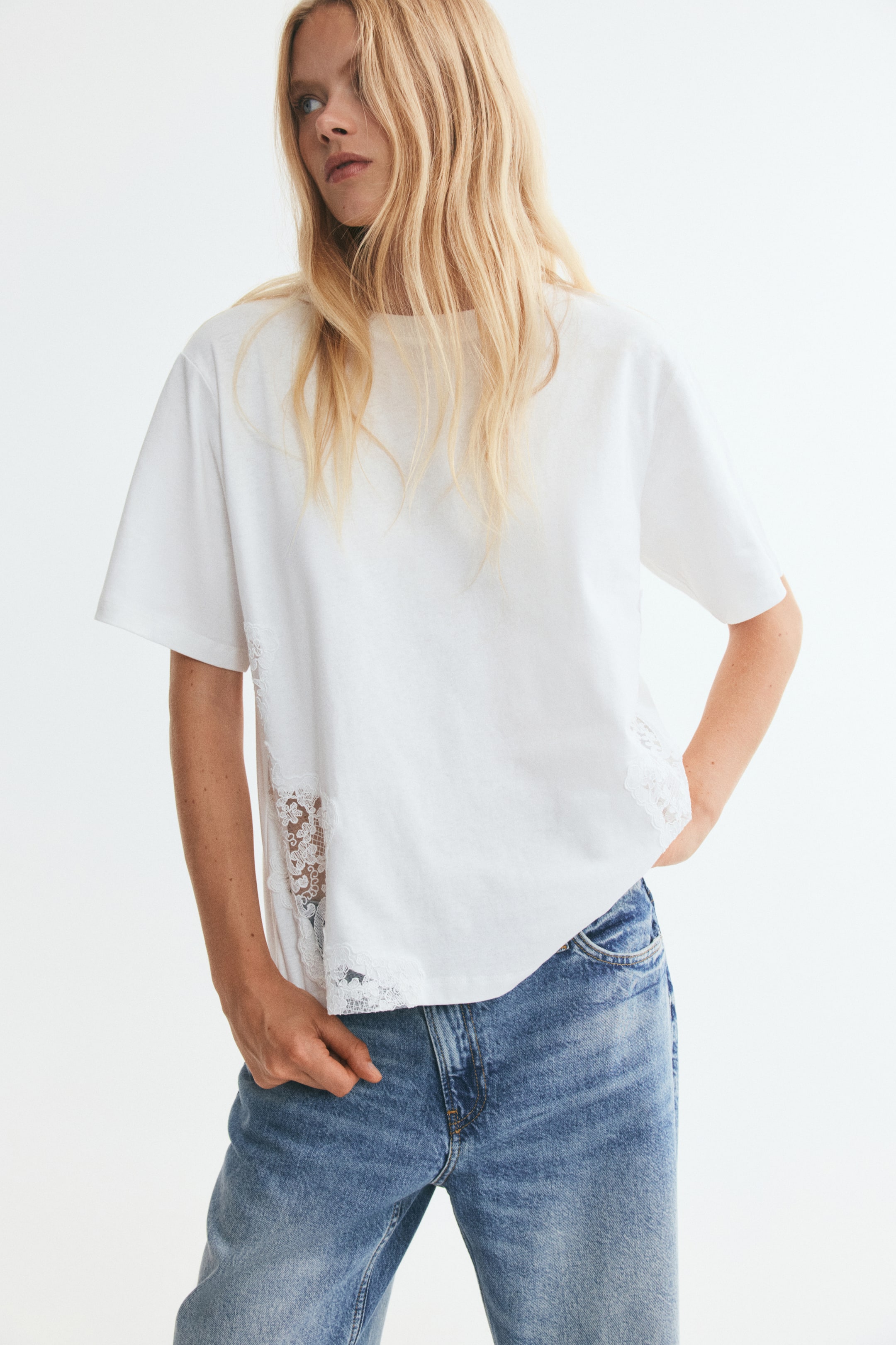 Vezi imaginea mai mare: Un tricou alb, cu croială lejeră, cu decolteu rotund, umeri căzuți și inserții delicate din dantelă de-a lungul părților inferioare este purtat de model. Modelul asortează tricoul cu blugi din denim albastru, sprijinind o mână aproape de buzunarul din față.