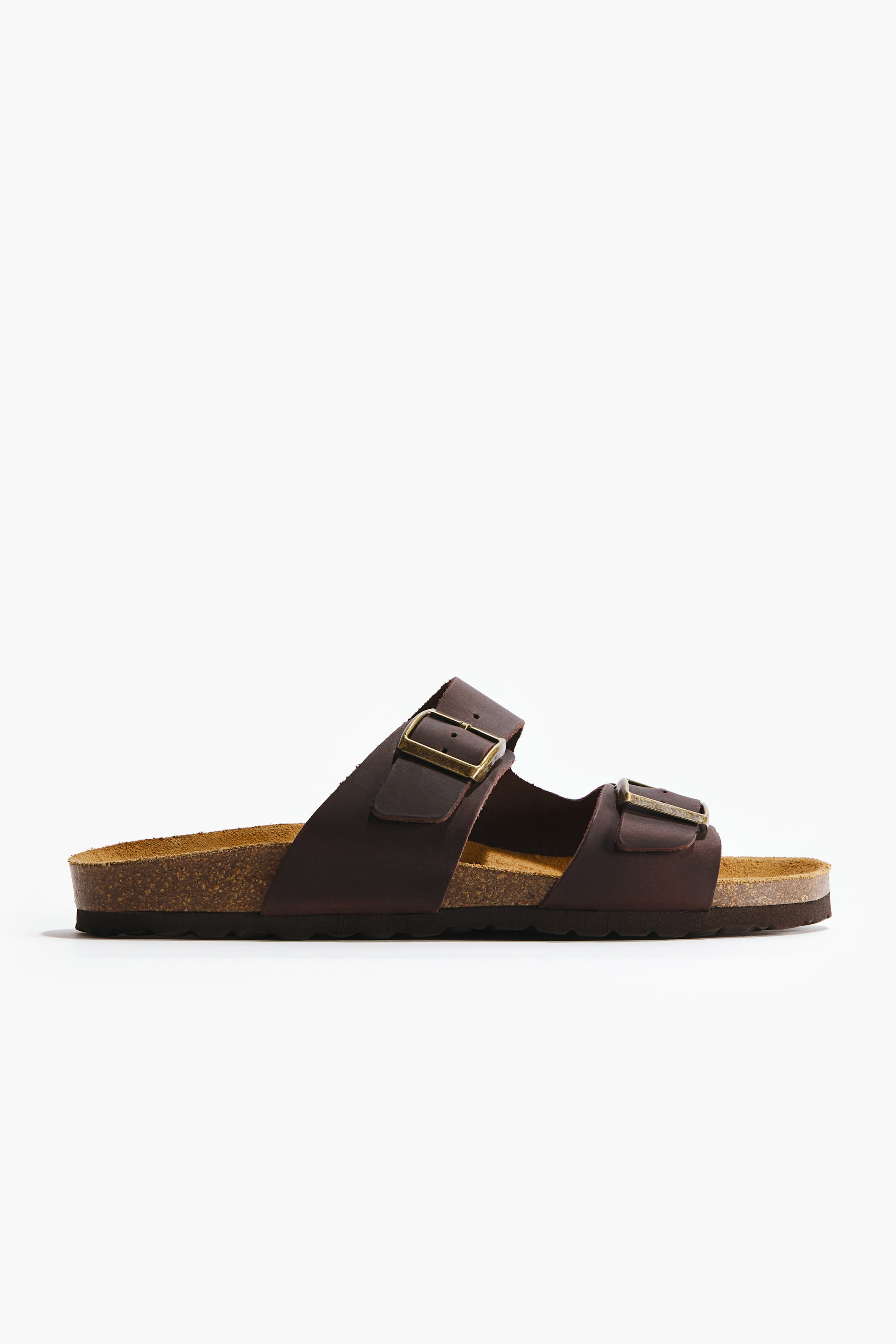View larger image: A dark brown sandal is visible from the side, featuring two wide straps with antique brass-coloured buckles. It showcases a light brown cork footbed and a dark brown sole.