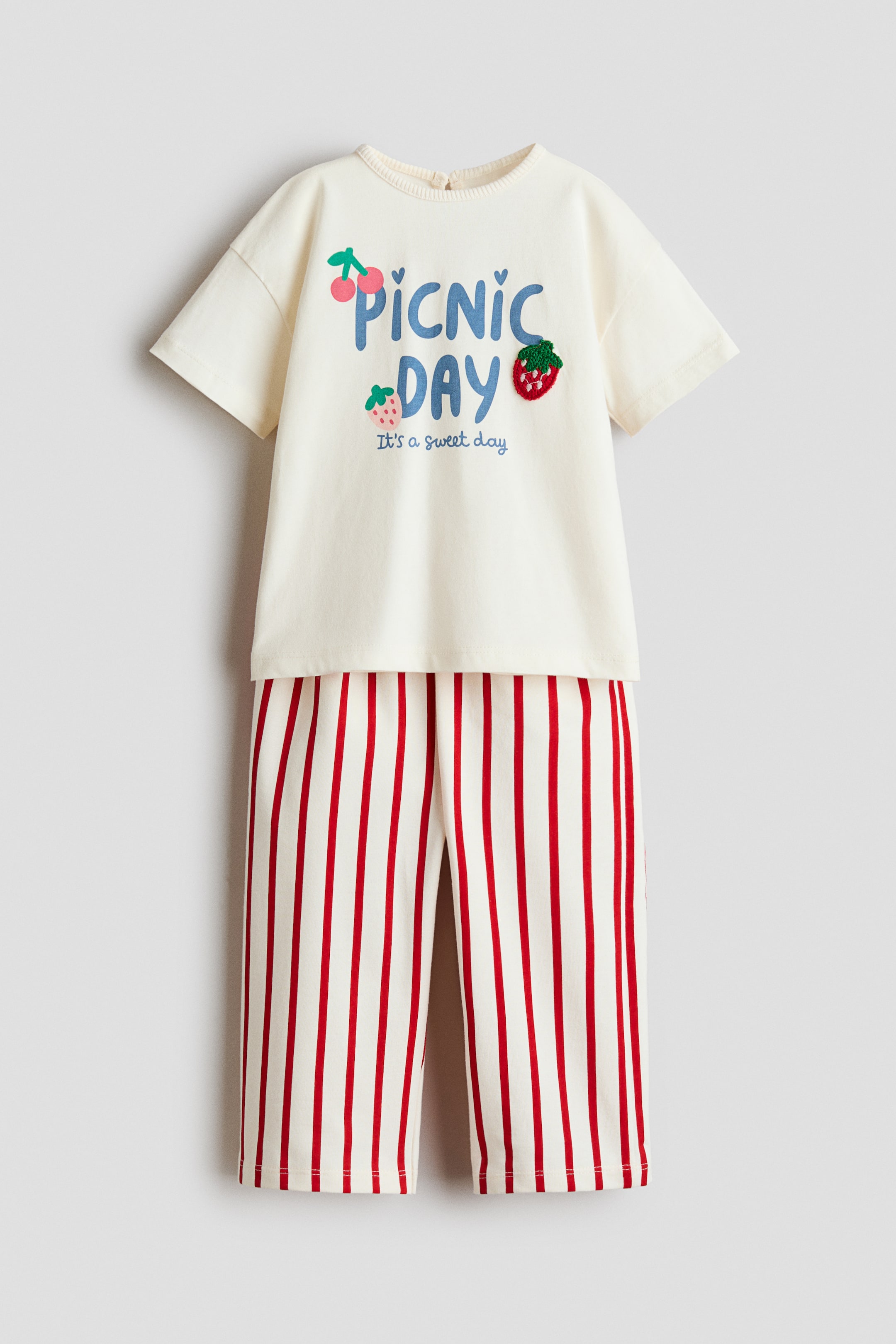 Grotere afbeelding bekijken: Een tweedelige kinderset toont een crèmekleurig T-shirt met korte mouwen en een bijpassende broek. Het T-shirt heeft een ronde hals, verlaagde schouders en op de voorkant een "PICNIC DAY"-opdruk met geborduurde aardbeien en kersen. De broek is ontworpen met een rechte pijp en verticale rode strepen.