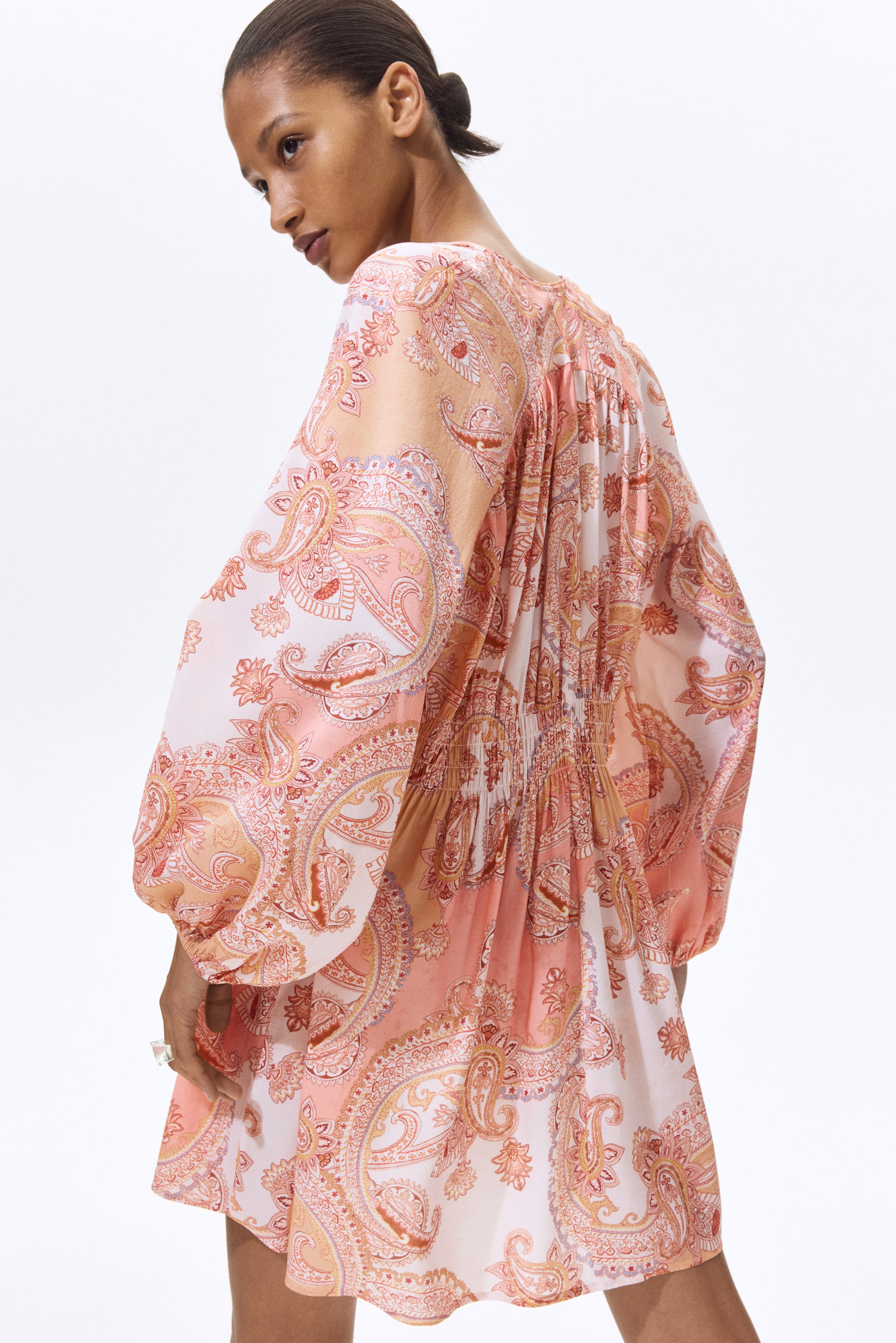 Agrandir l'image: Une robe courte, vue sous un angle arrière, affiche un imprimé cachemire dans des tons pêche, rose et blanc. Elle est dotée de manches longues volumineuses resserrées aux poignets et d'un panneau arrière froncé.